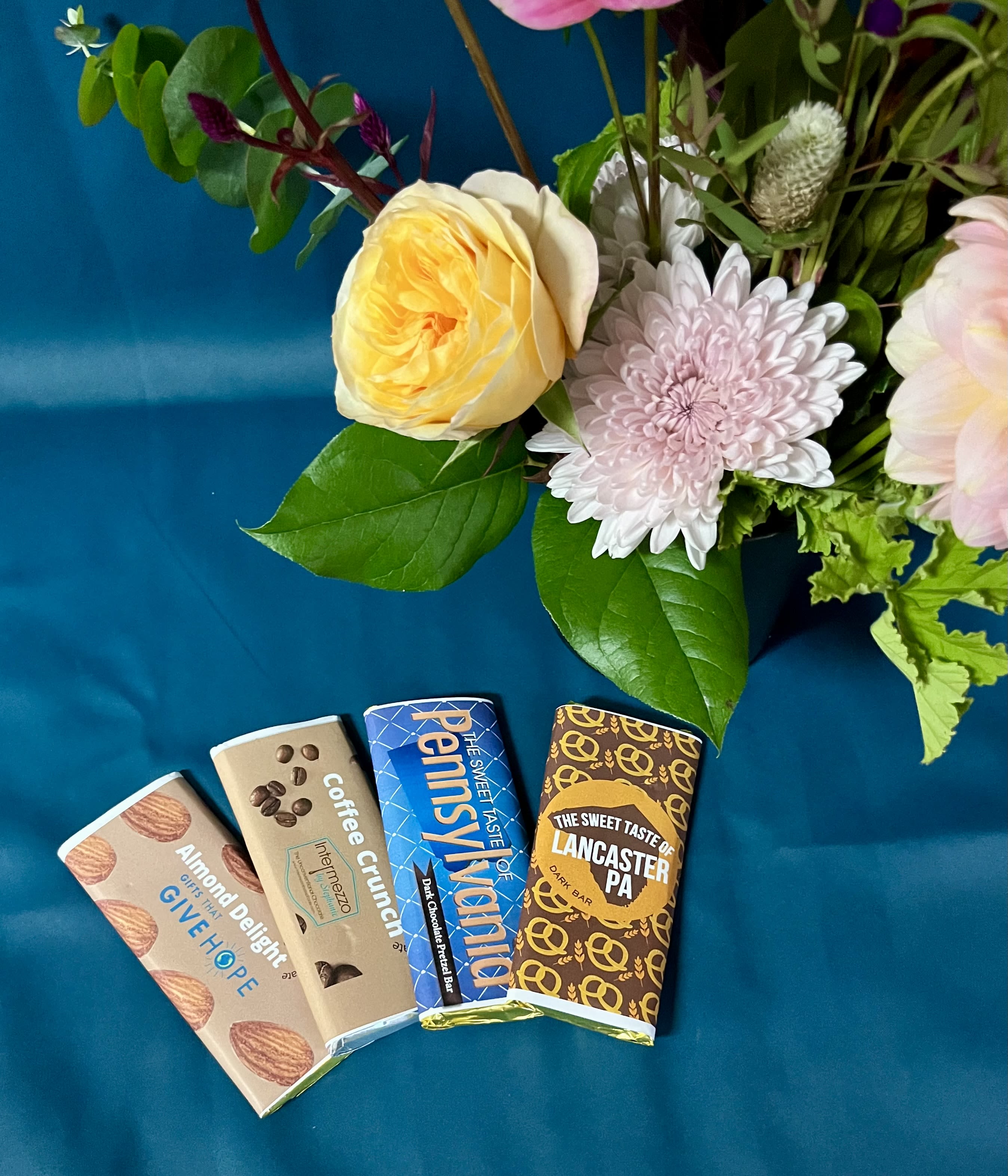Floral arrangement with pink and yellow blooms beside four wrapped chocolate bars on teal fabric.