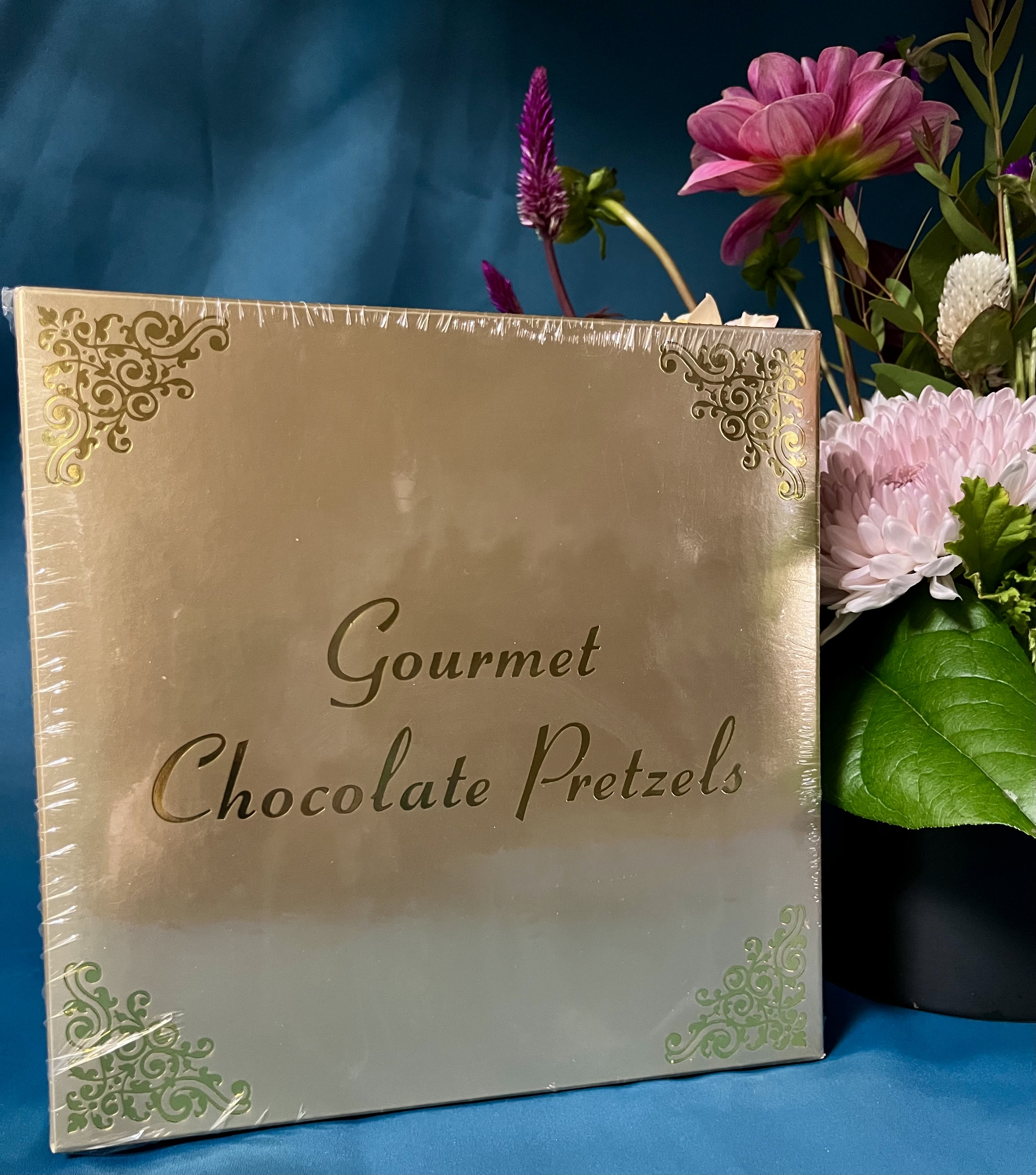 Gold box of gourmet chocolate pretzels beside a pink and purple flower arrangement on a teal background