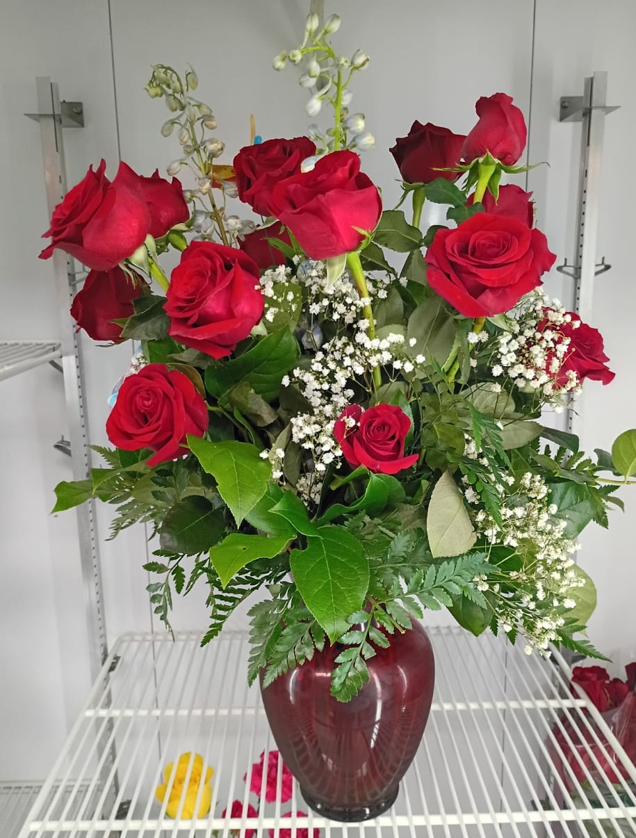 1 dozen red roses with mix greens and flowers - A cluster of a dozen deep red roses creates an immediate, romantic impression-perfect as an anniversary bouquet or a Valentine's Day gift that says "I love you" without words. The rounded burgundy glass vase and upright, full styling give the arrangement a classic silhouette so the person you're sending it to will feel cherished and remembered, whether marking a milestone or offering sympathy.  The bouquet pairs rich red roses with delicate white filler blooms and mixed greenery, including fern-like foliage, for a layered, textured look. Arranged at varying heights in a compact, rounded burgundy vase, it reads elegant from every angle and fits neatly on a bedside table or mantel. Vase included; care is simple-fresh water and a cool spot will keep the roses looking their best for days.