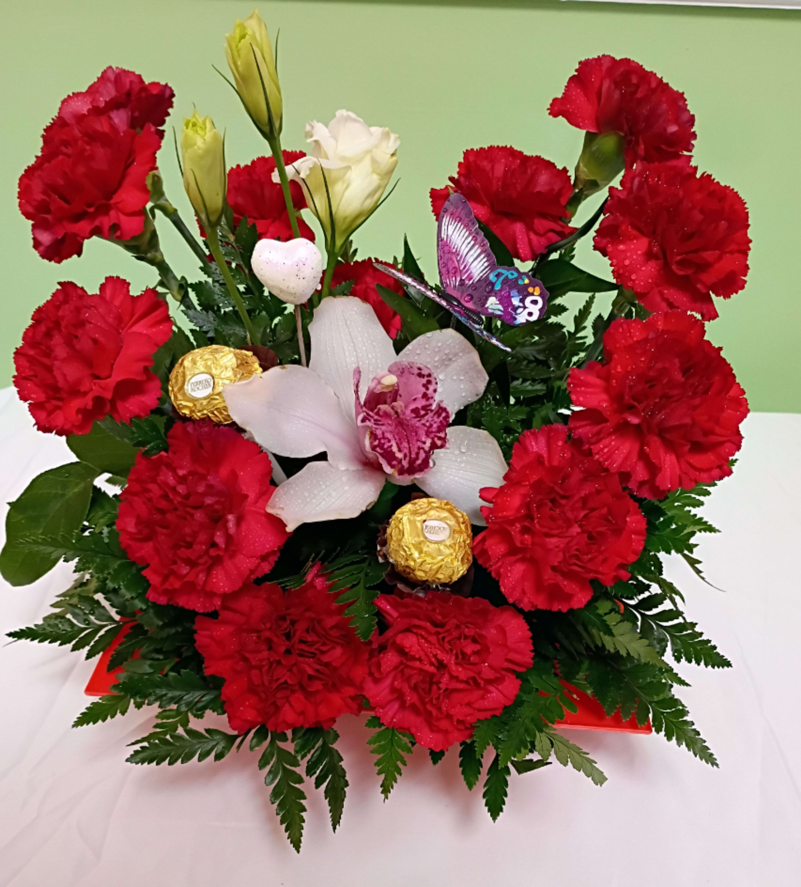 Carnation heart red with orchid and premium chocolate - A bold heart-shaped ring of bright red carnations frames a creamy cluster of white orchids in this romantic flower arrangement-perfect as a Valentine's Day or anniversary flower arrangement to make the person you're thinking of feel adored. The vivid red-and-white contrast reads festive and intimate, while the low, full tabletop silhouette and playful purple butterfly pick make it feel like a cherished gift basket meant to be displayed and admired.  This arrangement includes fragrant red carnations, white orchids and pale buds set on a red rectangular tray, accented with two gold-wrapped Ferrero Rocher chocolates and a purple butterfly decoration for an extra-special touch. The compact, dense construction sits low for easy placement on a dining table or desk and combines lush texture (the ruffled carnations and smooth orchid petals) with glossy hard-goods to create a luxurious yet approachable presentation.  A meaningful choice for anniversaries, Valentine's Day, or a heartfelt "I'm thinking of you" moment, this piece arrives ready to gift-no vase needed. For best enjoyment, keep orchids and carnations in a cool spot away from direct sun and replace water as needed to preserve freshness.