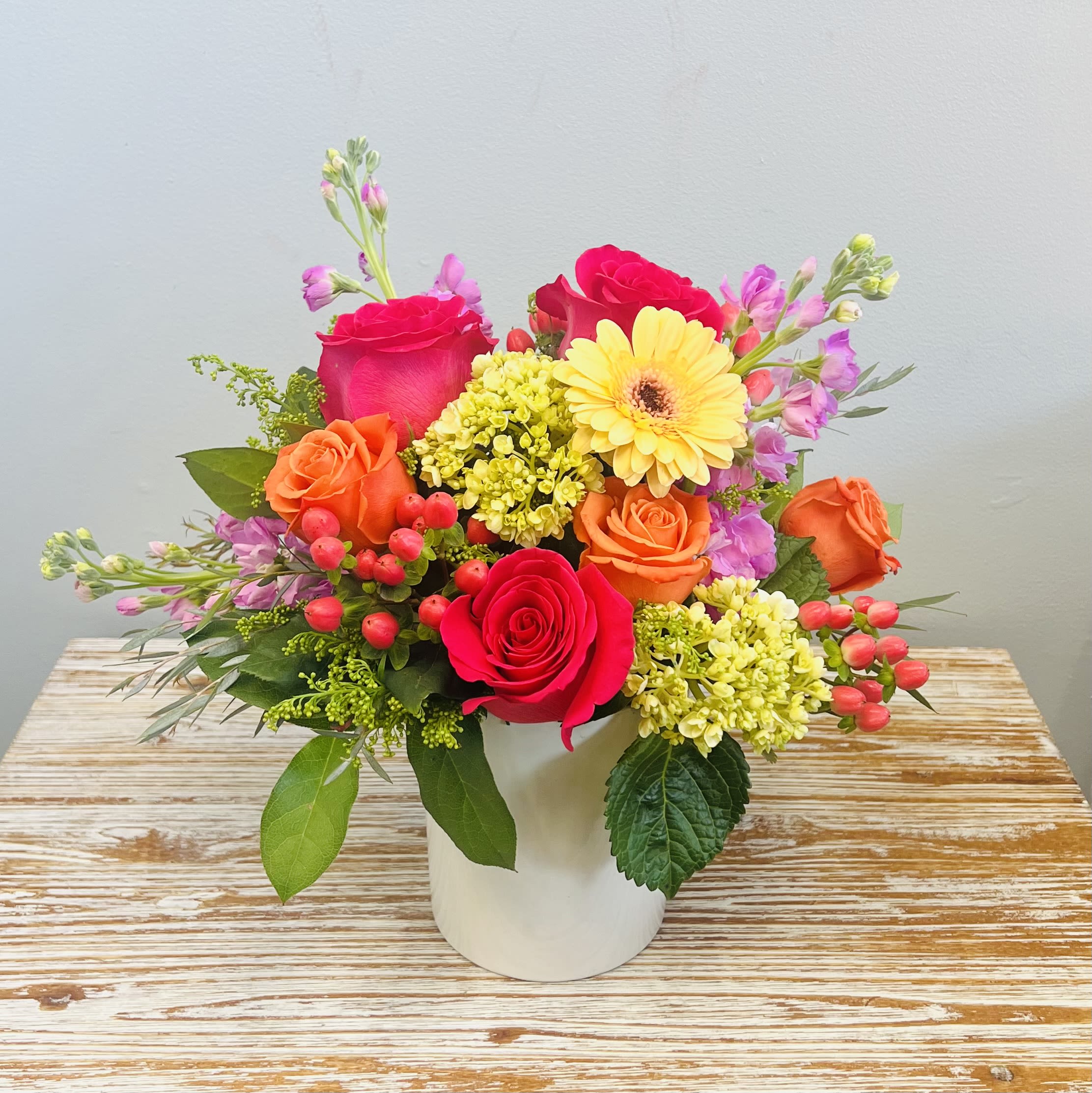 Cosmopolitan - Vivid hot pink and orange roses take center stage in this Cosmopolitan flower arrangement, a cheerful bouquet built to make Mom, or someone you love, feel celebrated. Perfect as a Mother's Day surprise or a bright Valentine's Day gift, the compact design balances bold color with a modern silhouette so the recipient immediately senses joy and warmth.  The arrangement pairs roses with tall pink stock stems for height, yellow gerbera daisies as a focal contrast, and clusters of green hydrangeas plus pink berry accents for texture and interest. Lush green foliage fills the rounded shape, and everything is gathered in a simple white ceramic cylinder vase that highlights the saturated pinks, oranges, and yellows for a clean, contemporary presentation.  At about 14" tall, Cosmopolitan fits nicely on a table or desk and arrives ready to display in its white ceramic vase. It's designed to brighten a birthday, say "I'm thinking of you," or add a vivid pop of color to holiday moments.