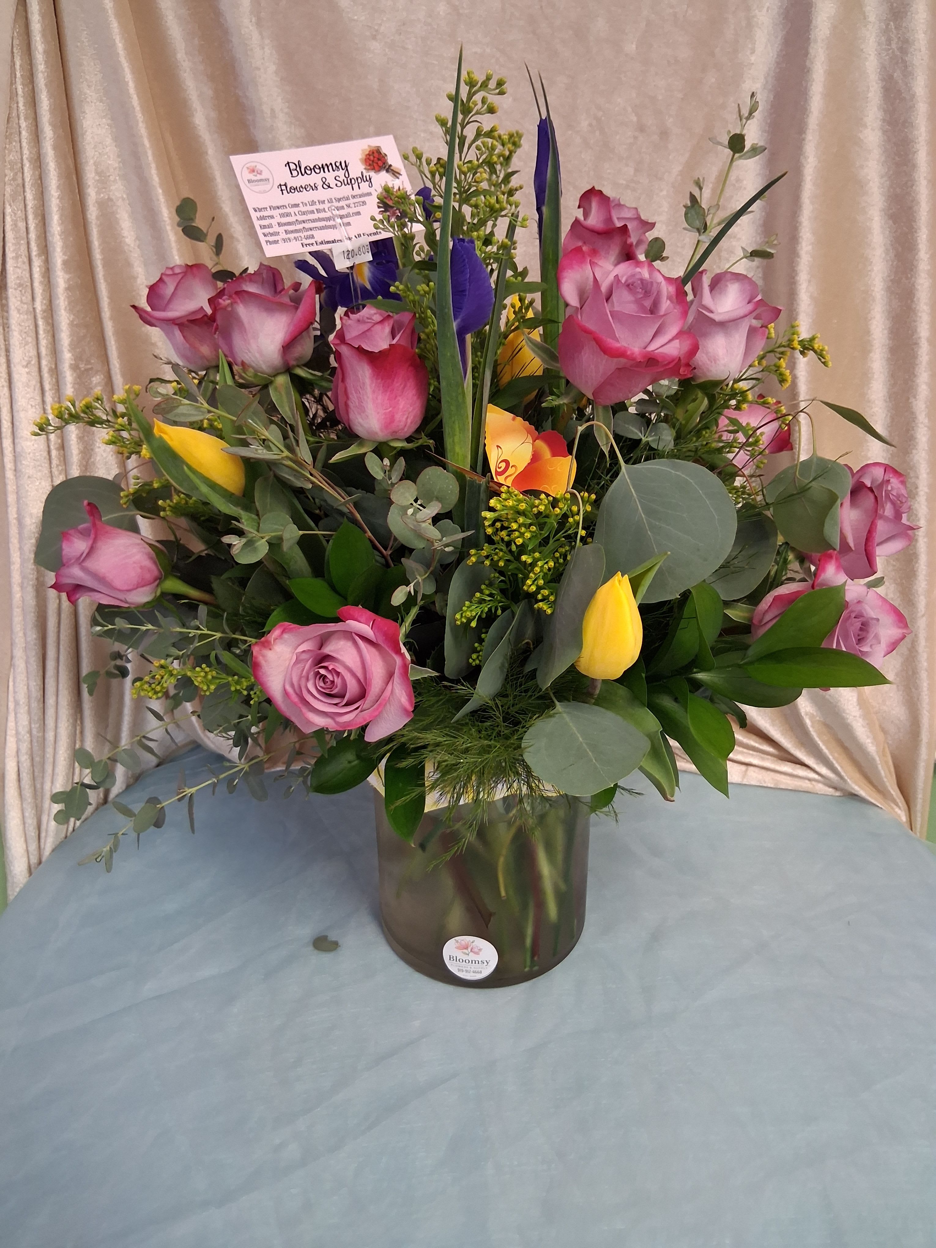  20 purple roses with tulips and mixed greens - A bright, romantic bouquet built around one dozen purple-accented roses makes a cheerful birthday or thank-you gift that feels both polished and personal. This flower arrangement for spring celebrations pairs rounded pink roses with sunny yellow tulips and tall purple accent blooms to create an uplifting color story that reads joyful from across the room. The arrangement is presented in a clear glass vase and arrives with a floral pick card ready for your message.  The design is full and upright, layered with the soft texture of rose heads, the smooth, cupped shapes of tulip buds, and airy greenery-broad eucalyptus-like foliage that adds cool green depth and a graceful silhouette. Colors include pink, yellow, purple, and rich green; stems are arranged to showcase contrasting heights and textures for instant visual impact. Sized to sit on a table or desk, this bouquet is the kind of gift that makes the person you're thinking of feel seen and celebrated.