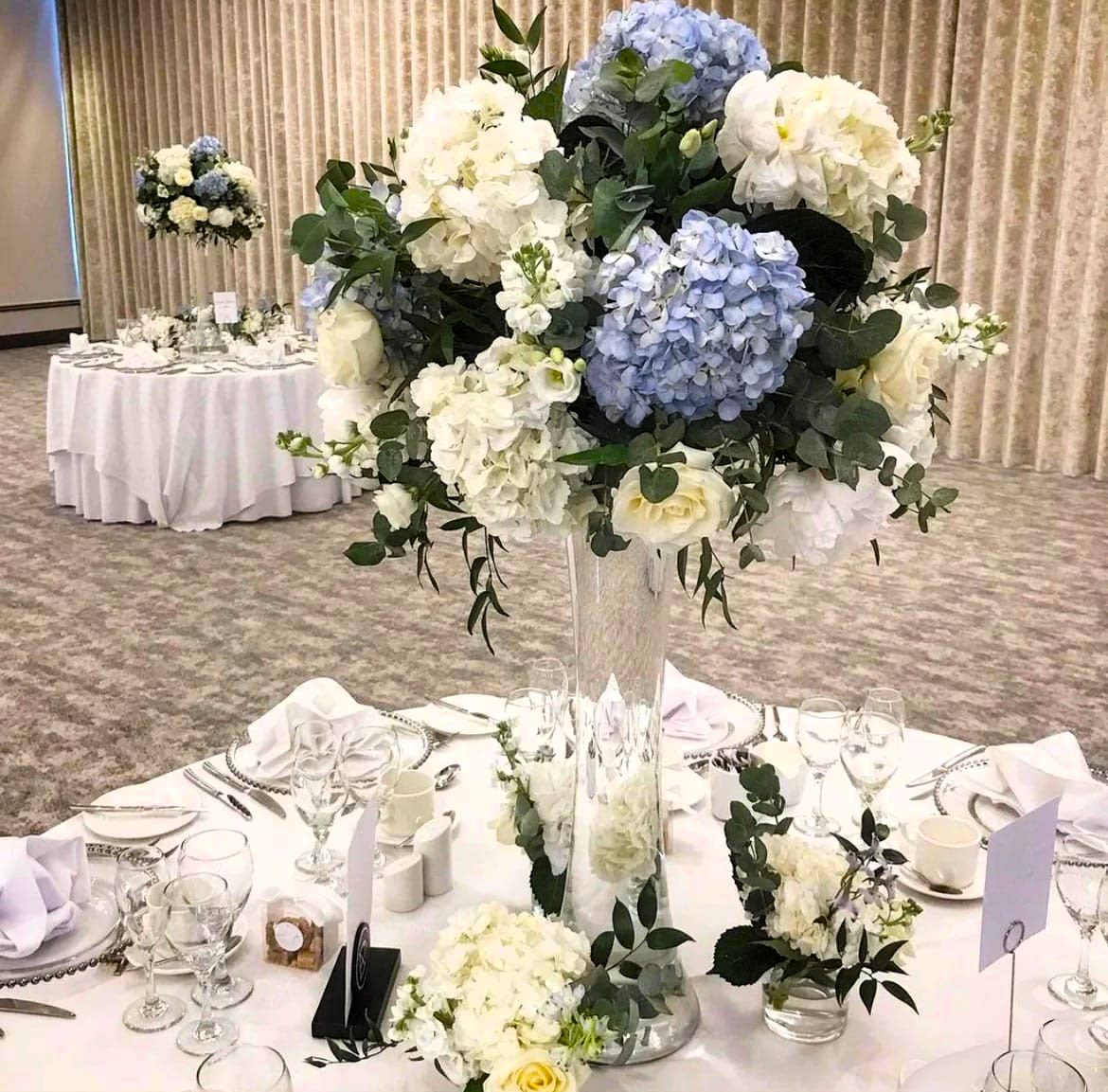 Table arrangement blue and white for special events  - Pale blue and white hydrangeas form the heart of this tall, formal flower arrangement-an elegant wedding or special events centerpiece designed to make the person you're honoring feel truly celebrated. A rounded silhouette of large hydrangea clusters is accented with classic white roses and wisps of airy greenery, creating a soft, refined look that reads both romantic and effortlessly sophisticated for anniversaries, rehearsal dinners, or upscale corporate receptions.  Arranged in a clear glass vase to keep the composition light and luminous, the bouquet's white, blue, and green palette complements dressed banquet tables and coordinated place settings. The full, rounded flower masses provide visual impact from every angle without blocking conversation, and the added table number card and matched dinnerware, glassware, and napkins in the display show how easily this centerpiece integrates into a curated event design.  Perfect for couples seeking a timeless floral statement or hosts planning a polished celebration, this arrangement brings floral texture, gentle color, and a graceful height to your table. For event orders, consider the table dimensions and sightlines-we recommend confirming quantity and placement with our team to ensure consistent coverage and the best presentation throughout your reception.