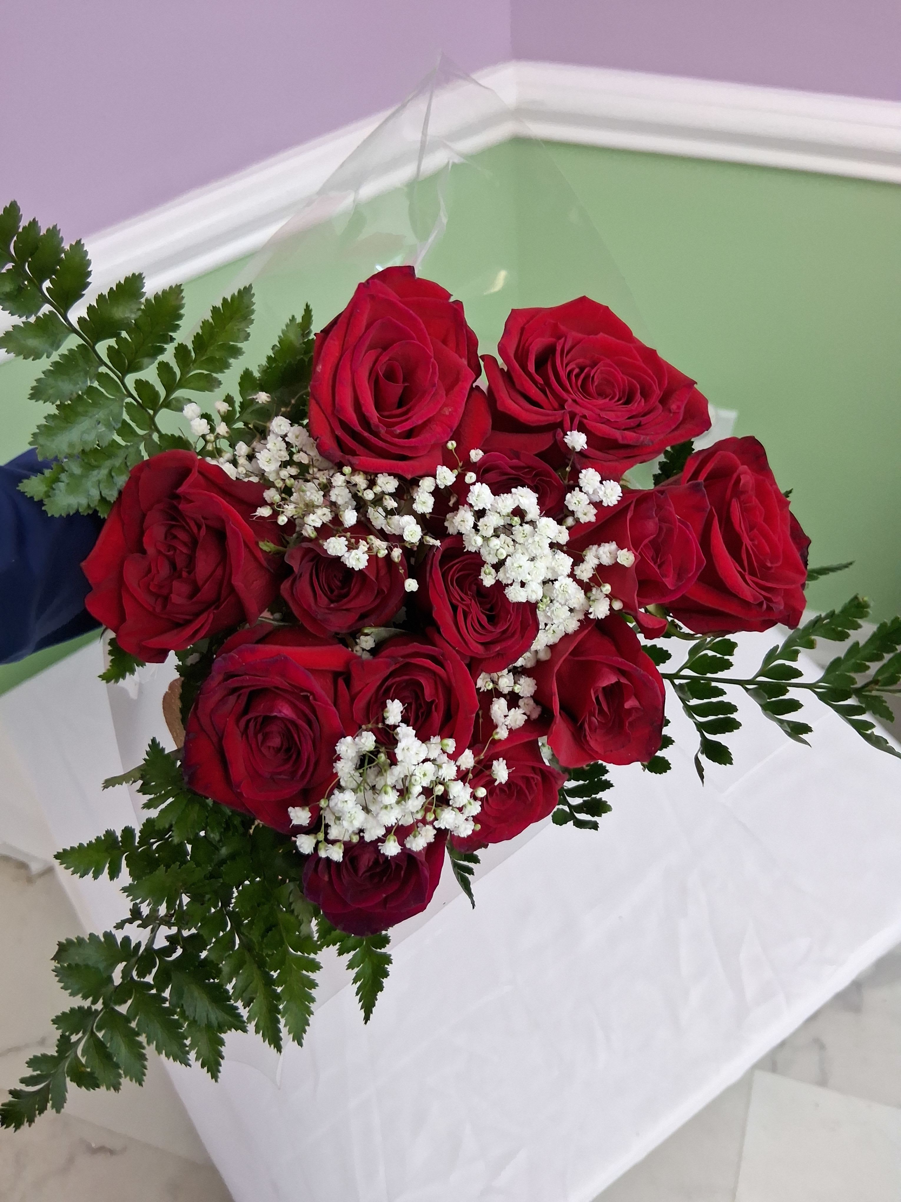 One dozen classic red roses bouquet - A dense cluster of deep red roses makes an immediate romantic statement-perfect as a red roses anniversary bouquet or a Valentine's Day gift that says "I love you" without words. The arrangement reads classic and timeless, with the recipient feeling adored the moment they see the rich crimson blooms framed by airy greenery.  Twelve long-stemmed red roses are neatly gathered with delicate white baby's breath and feathery fern foliage for texture and contrast. The stems are finished in clear plastic wrap and tied with a simple white ribbon for a fresh, hand-tied presentation that keeps the focus on the flowers. The red, white, and green palette is traditional and elegant, suitable for romantic celebrations or to mark a special milestone.  This arrangement is ideal for anniversaries, romantic gestures, or to make someone feel truly cherished on a special day. For best enjoyment, recut stems and place the bouquet in fresh water on arrival; sizing and stem length match standard dozen-rose bouquets for easy display in most vases.
