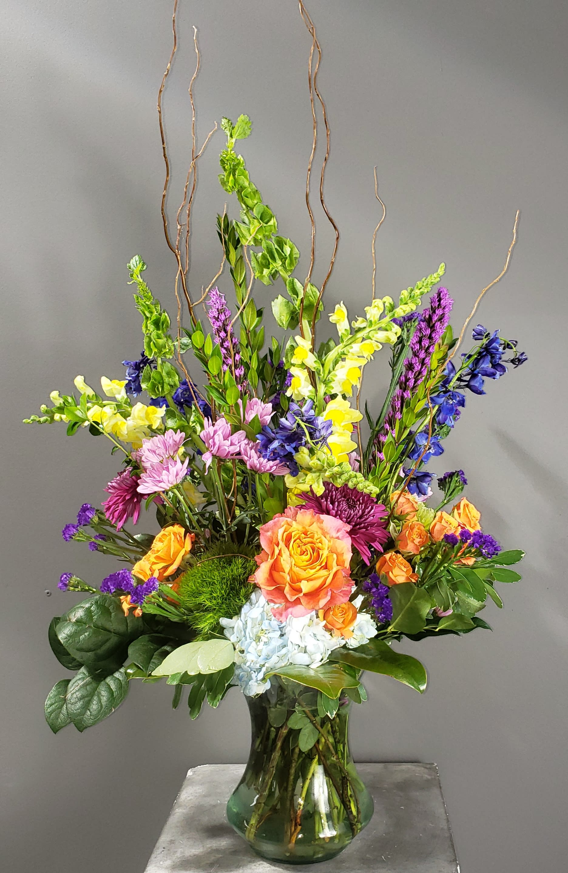 Tall mixed bouquet in a glass vase with orange, yellow, purple, and pink flowers