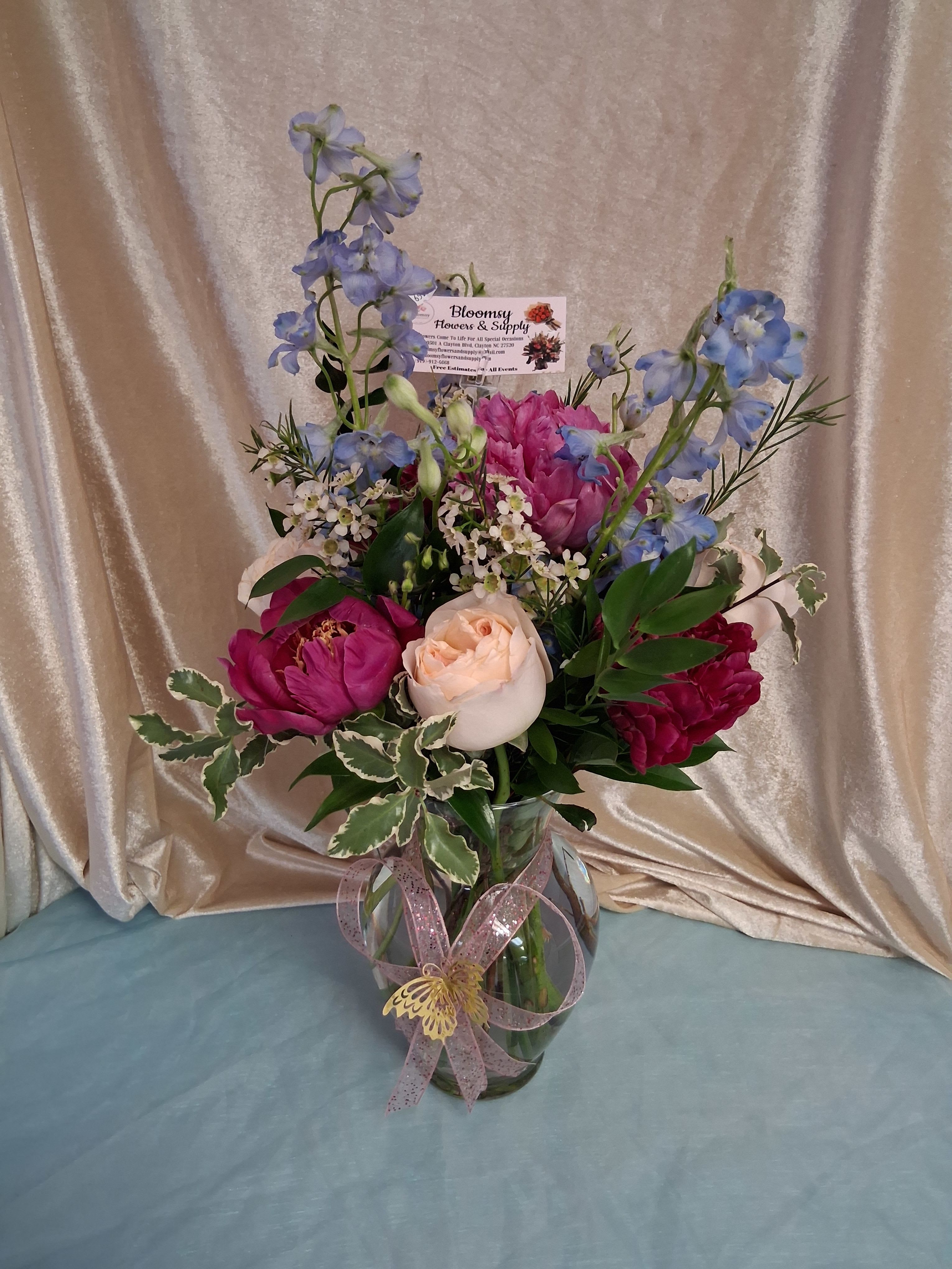 Peonies in glass vase - Lush pink peonies and soft peach roses combine into a romantic birthday bouquet or anniversary arrangement that feels both celebratory and intimate. The tall blue delphinium spikes add elegant height above the dense cluster of pink blooms, while touches of white filler flowers and variegated foliage create a light, airy contrast-an arrangement meant to make the person you're thinking of feel truly seen and celebrated.  Carefully arranged in a clear glass vase and finished with a pink ribbon, this vase arrangement arrives gift-ready and polished; a decorative butterfly pick and a floral business card add a thoughtful, playful touch. The composition is upright and full, with a mix of large peony-like blooms, roses, delphinium, and small white accents that offer visual texture and gentle movement-perfect for birthdays, anniversaries, thank-you gestures, or simply brightening someone's day.