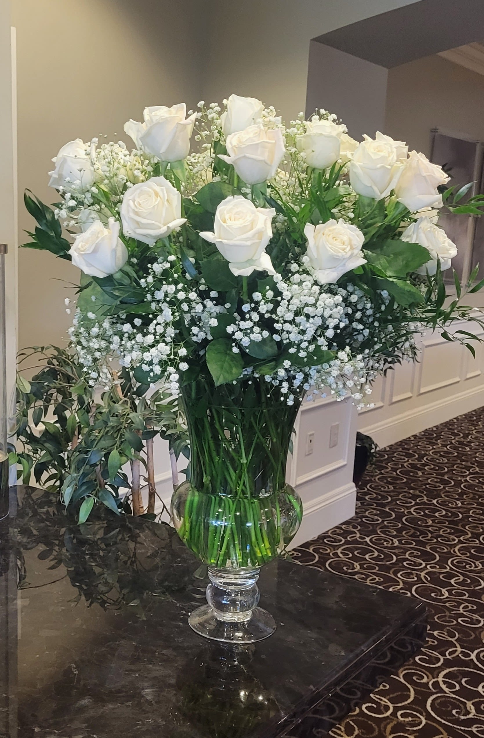 Two dozen classic white roses in vase  - A cloud of two dozen classic white roses gathered with delicate baby's breath creates a serene, timeless gift-perfect as a sympathy arrangement or an elegant wedding bouquet displayed on a reception table. The clean white-and-green palette and upright, symmetrical shape give the recipient a sense of calm and reverence, the kind of arrangement that makes someone feel truly honored and supported.  Each stem is arranged in a clear glass vase so the long green stems and fresh water are visible, reinforcing the arrangement's crisp, formal look. The composition is full and balanced: 24 white roses nestled among airy sprays of baby's breath for soft texture and gentle contrast. The vase's simple clarity keeps the focus on the blooms and complements both modern and traditional settings.  This is an ideal choice when you want to convey sympathy, send wedding wishes, or mark an anniversary with understated dignity-the kind of bouquet that reads as thoughtful and respectful. Vase-included; standard care applies (fresh water, trim stems every few days) to keep the roses looking their best for as long as possible.