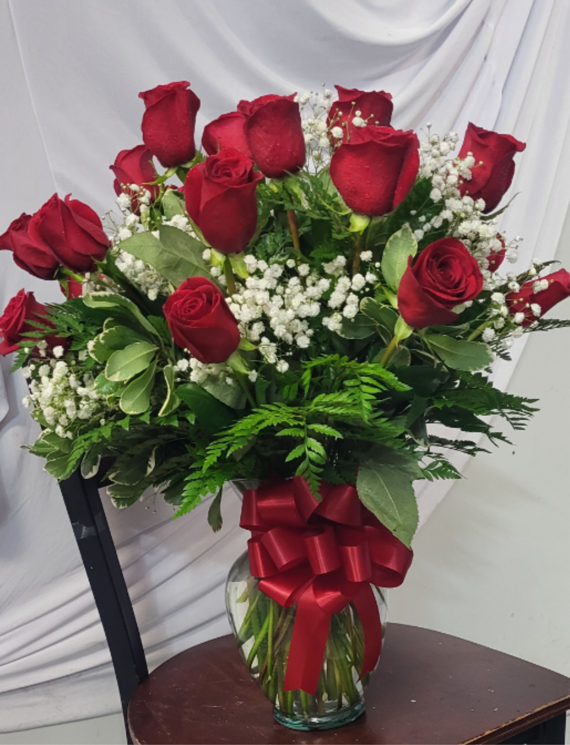 Classic two dozen red roses in vase - A lush cluster of two dozen deep red roses arranged with delicate sprays of white baby's breath creates an unmistakably romantic birthday bouquet, anniversary roses arrangement, or Valentine's Day gift that reads classic and heartfelt. The rich crimson blooms sit upright in a clear glass vase, tied with a large red satin ribbon, so the person you're thinking of will feel seen and cherished the moment they walk in the room.  This formal, full composition pairs glossy green foliage with the soft texture of baby's breath for striking red-and-white contrast that fits both celebratory and more solemn moments like sympathy. The arrangement ships in a clear glass vase finished with a red ribbon-ready to display on a table or mantle-and is sized as a traditional two dozen-rose presentation for a generous, show-stopping presence.