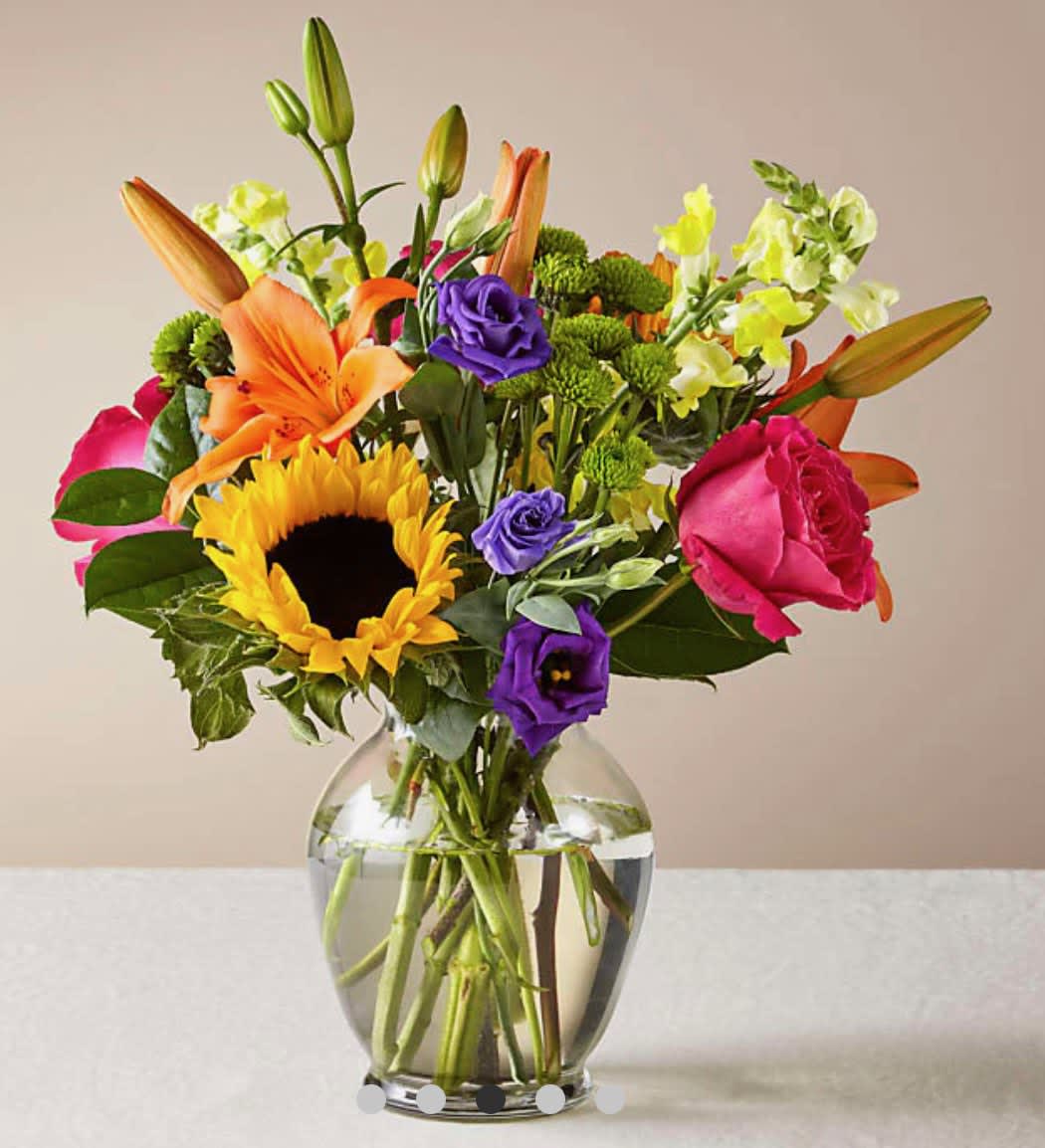 Colorful mixed bouquet in a clear glass vase with a sunflower, roses, lilies, and purple blooms