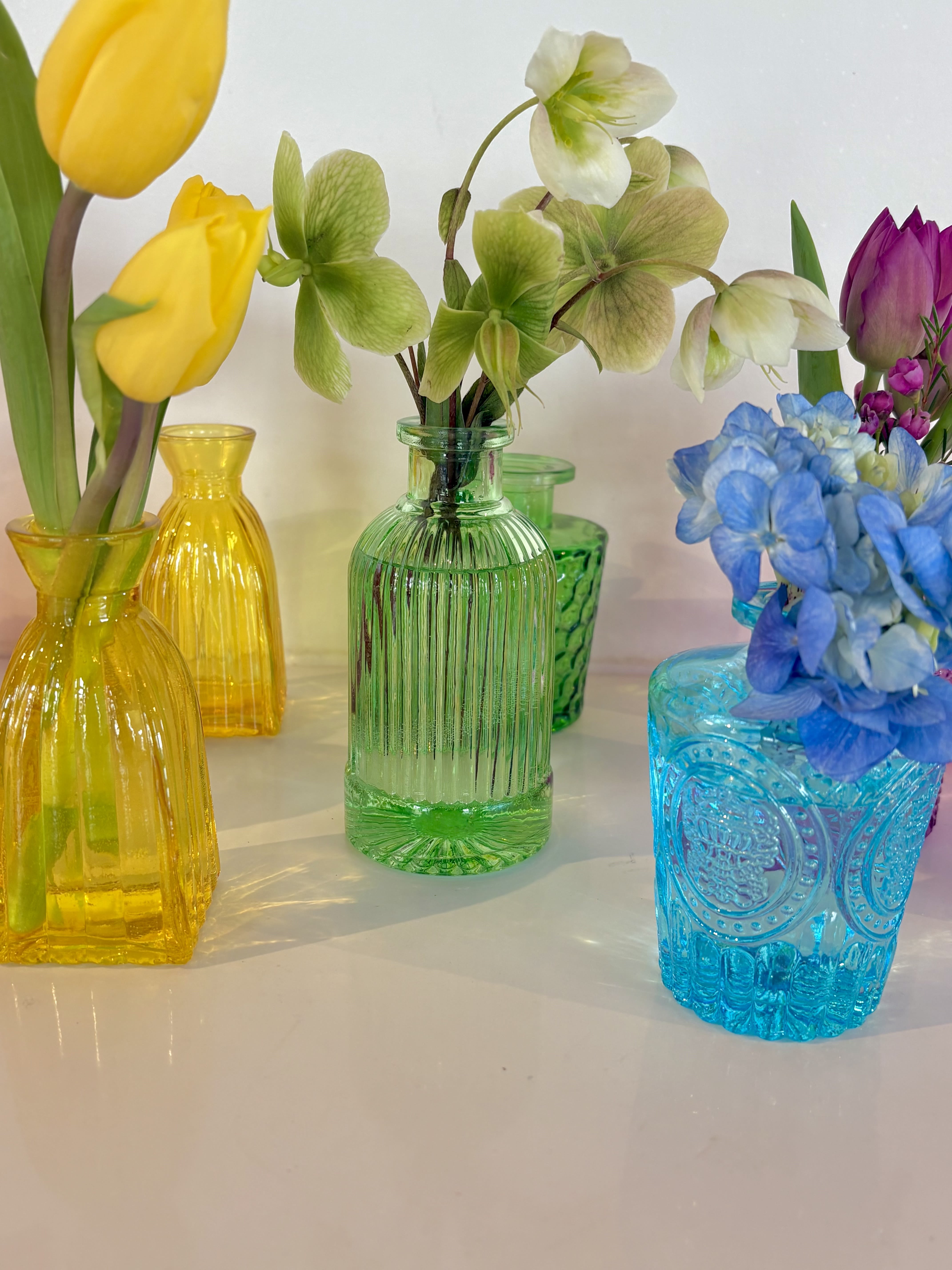 Bud Vases in Colors!  - A playful rainbow of petite bud vases brings instant cheer - perfect as a springtime centerpiece, party decor, birthday surprise, or an everyday "thinking of you" gift. This flower arrangement of mixed-color bud vases (yellow, blue, green, pink, purple, red) comes pre filled with seasonal blooms.   The translucent, patterned glass catches light and casts jewel-toned reflections.   Choose from standard size- 1 bud vases you select color, deluxe- 3 bud vases, premium- set of 6. You can select your colors.  We will let you know if they are not in stock.  