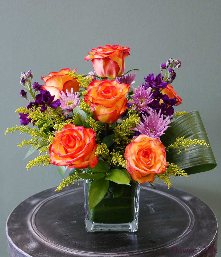 Bright arrangement of orange roses and purple flowers in a glass cube vase with a folded green leaf