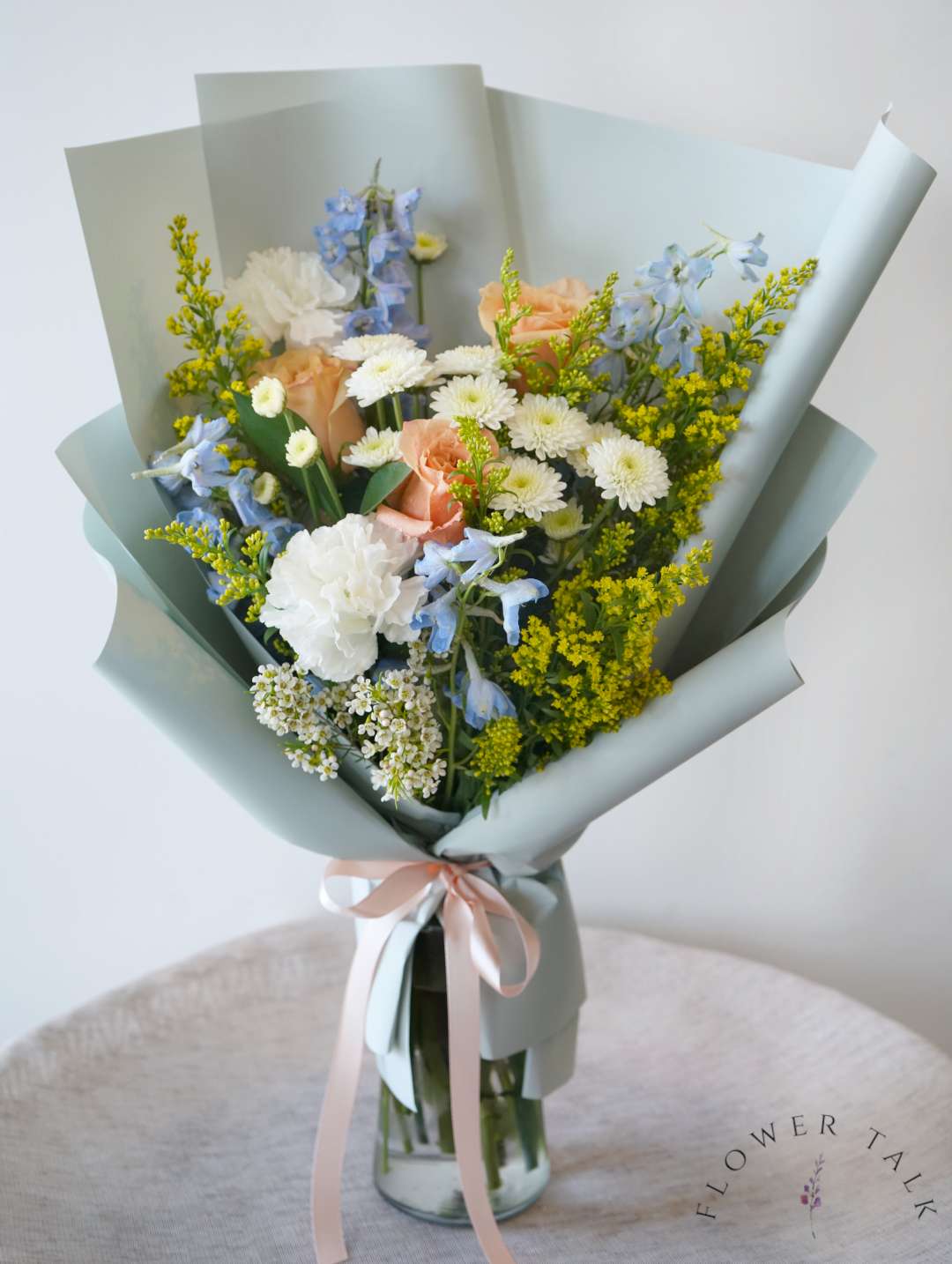 Bouquet of peach roses, white daisies, and blue flowers wrapped in pale paper in a glass vase