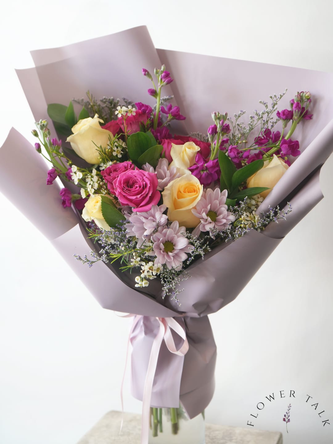 Hand-tied bouquet of yellow and pink roses with purple flowers wrapped in lavender paper.