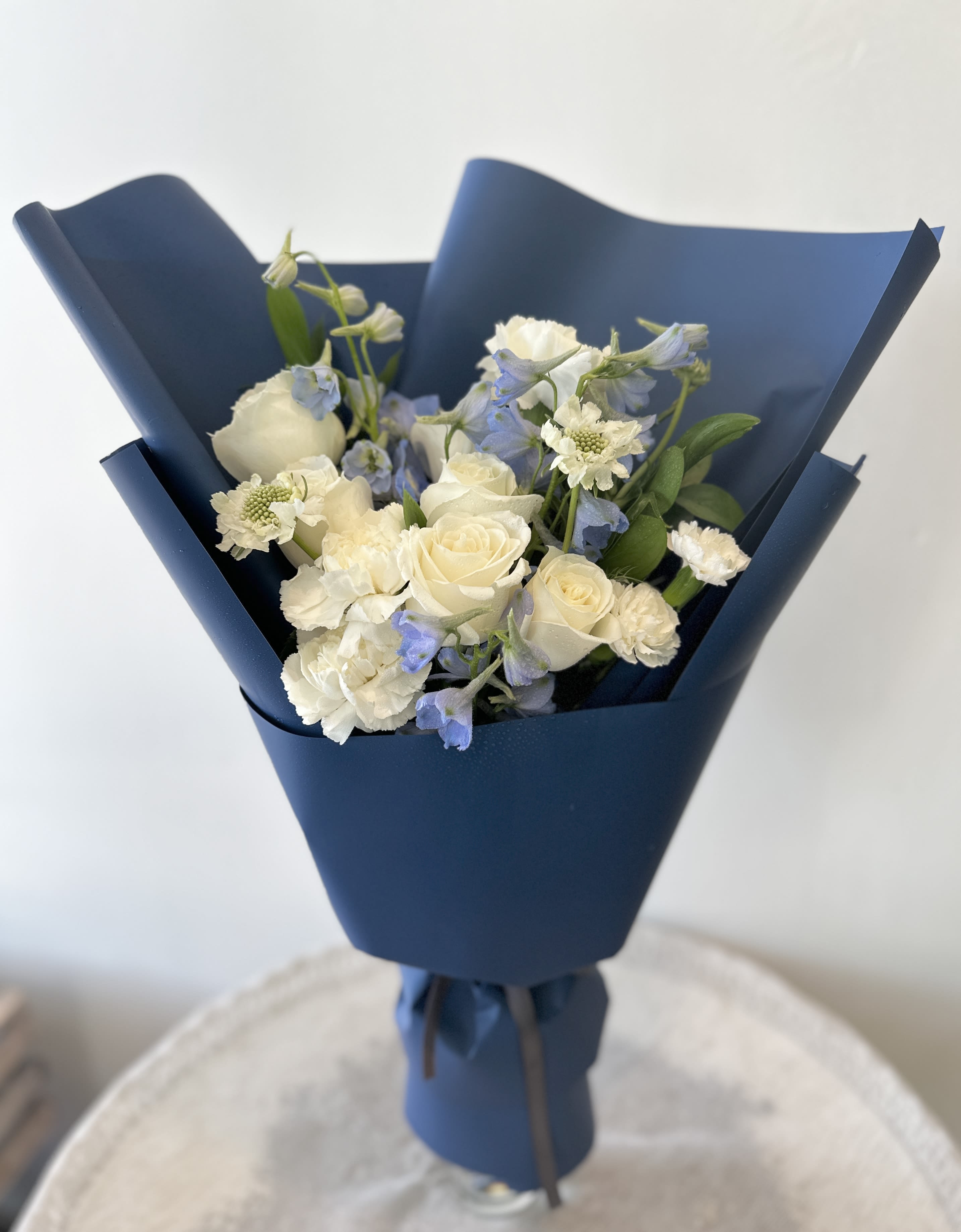 Blue Night Sky Hand Bouquet - Creamy white roses mingle with delicate white blooms and pale blue delphinium in this serene hand-tied flower arrangement - a Blue Night Sky hand bouquet perfect for a thoughtful gift, spring celebration, or wedding-adjacent moment. The cool blue-and-white palette and soft textures give the recipient a sense of calm elegance; she'll feel quietly celebrated and seen by this understated, romantic presentation.  Note: Vase is NOT included and wrapping paper can change.