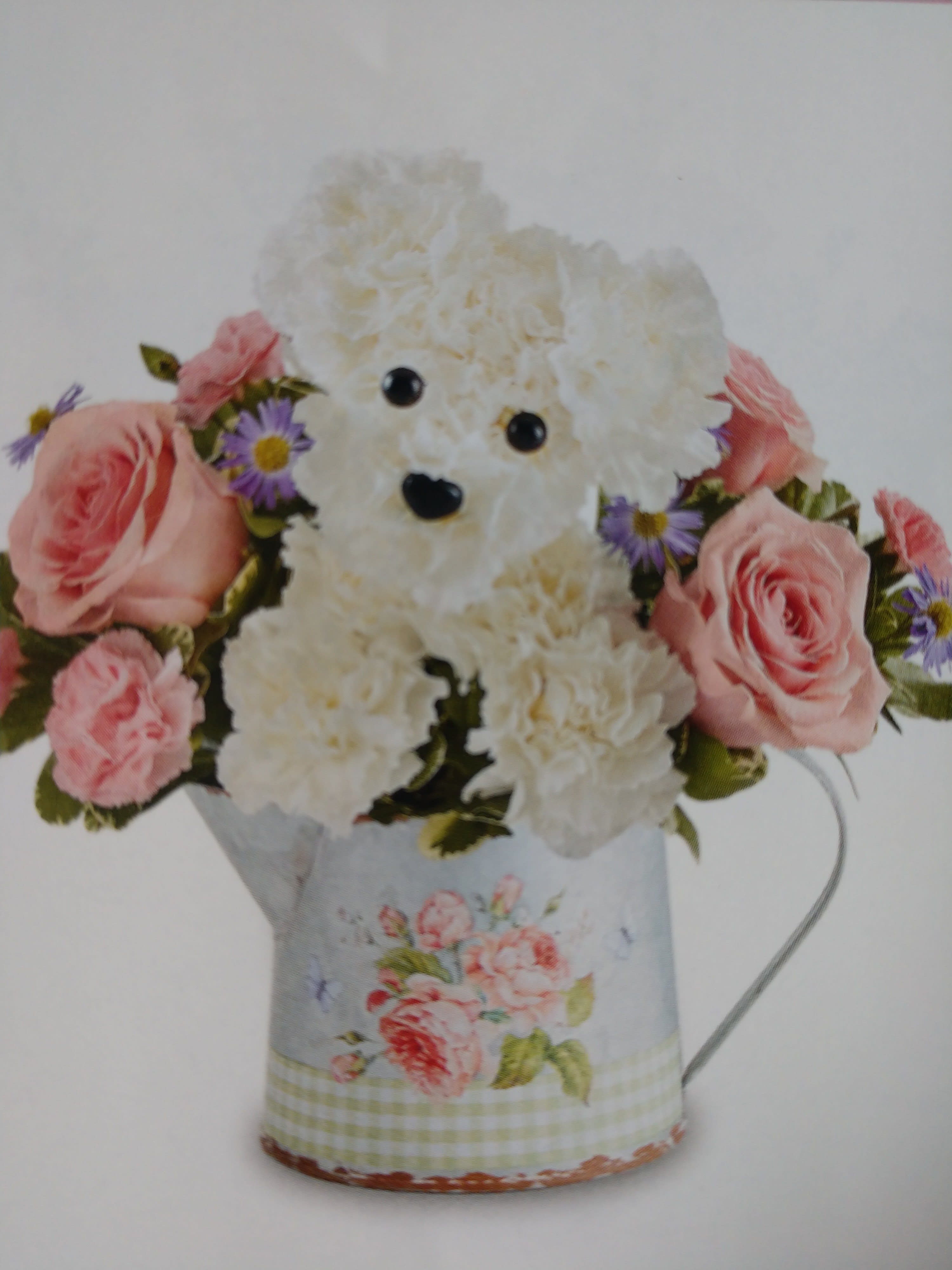 White teddy bear surrounded by pink roses and purple flowers in a floral tin