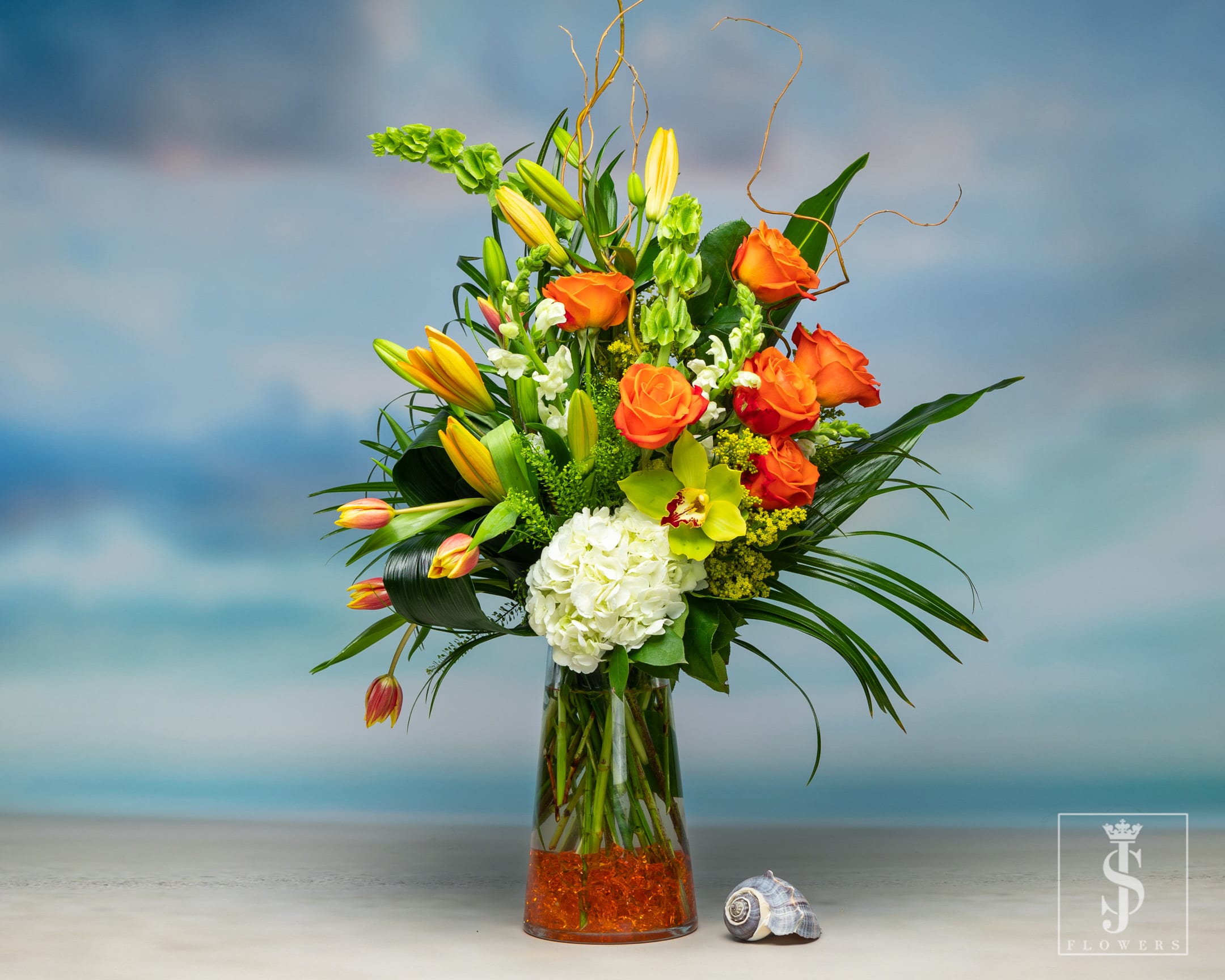 Citrus Summer Blossom - Citrus Summer Blossom is an elegant arrangement with Bells of Ireland, Lilies, Roses, and Hydrangeas, Solidago, upscale greens, and Orchids in a tall vase with Orange crystals for aesthetic flaire. 