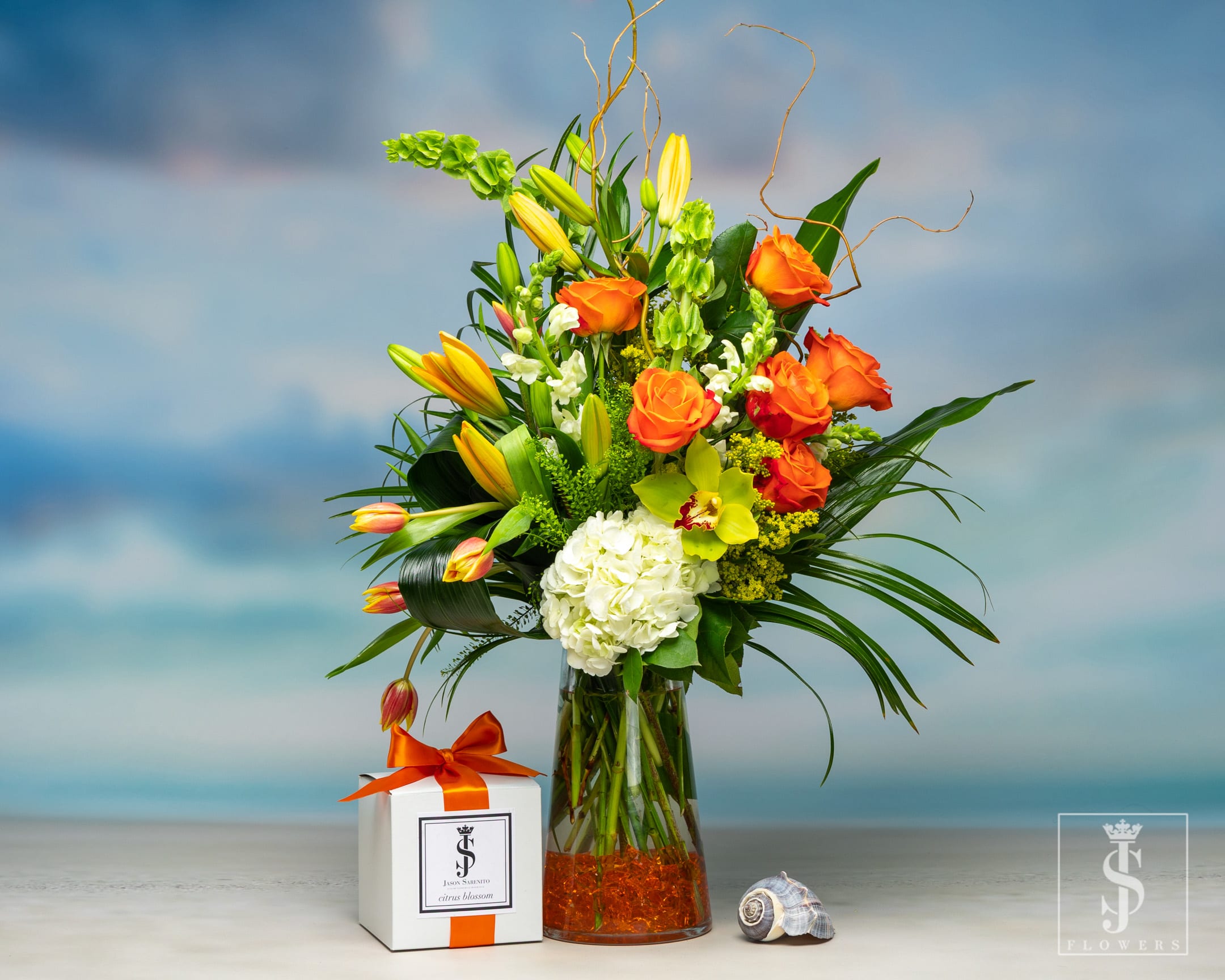 Citrus Summer Blossom with Citrus Blossom Candle - Citrus Summer Blossom Citrus Summer Blossom is an elegant arrangement with Bells of Ireland, Lilies, Roses, and Hydrangeas, Solidago, upscale greens, and Orchids in a tall vase with Orange crystals for aesthetic flaire. This comes paired with our Citrus Blossom Single Wick Candle. It is a delectable smell of flowering citrus blossom, lemongrass, orchard leaves, with undertones of fresh verbena, hints of gardenia, white lilac, lily of the valley, and clementine flower. 