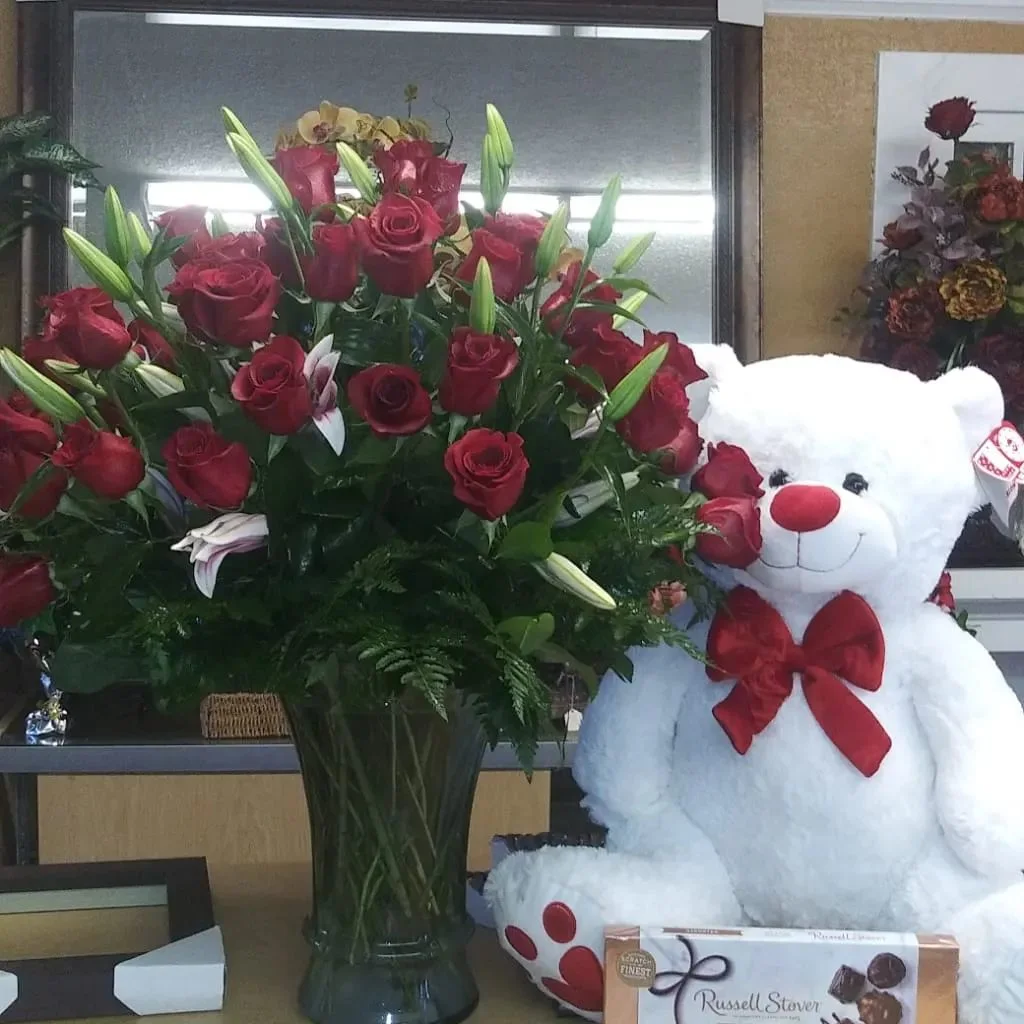Red roses and lilies in a glass vase beside a white teddy bear with a red bow