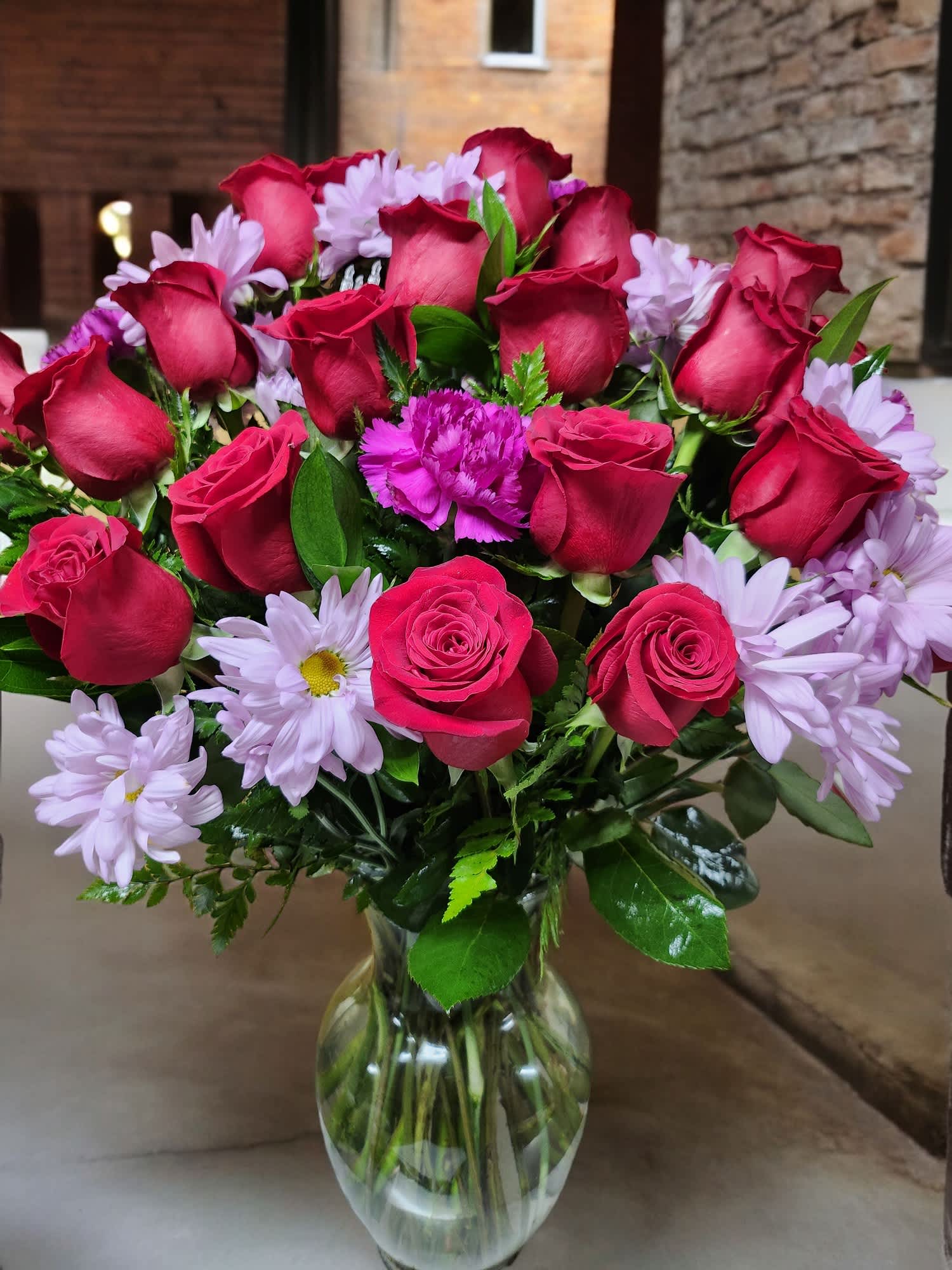 Roses for Daisy - Elevate your home with timeless elegance arrangement, featuring striking 18 red roses, carnation and lavender daisies in a glass vase. Perfect for expressing love and remembrance. Approximately 20w x 26h