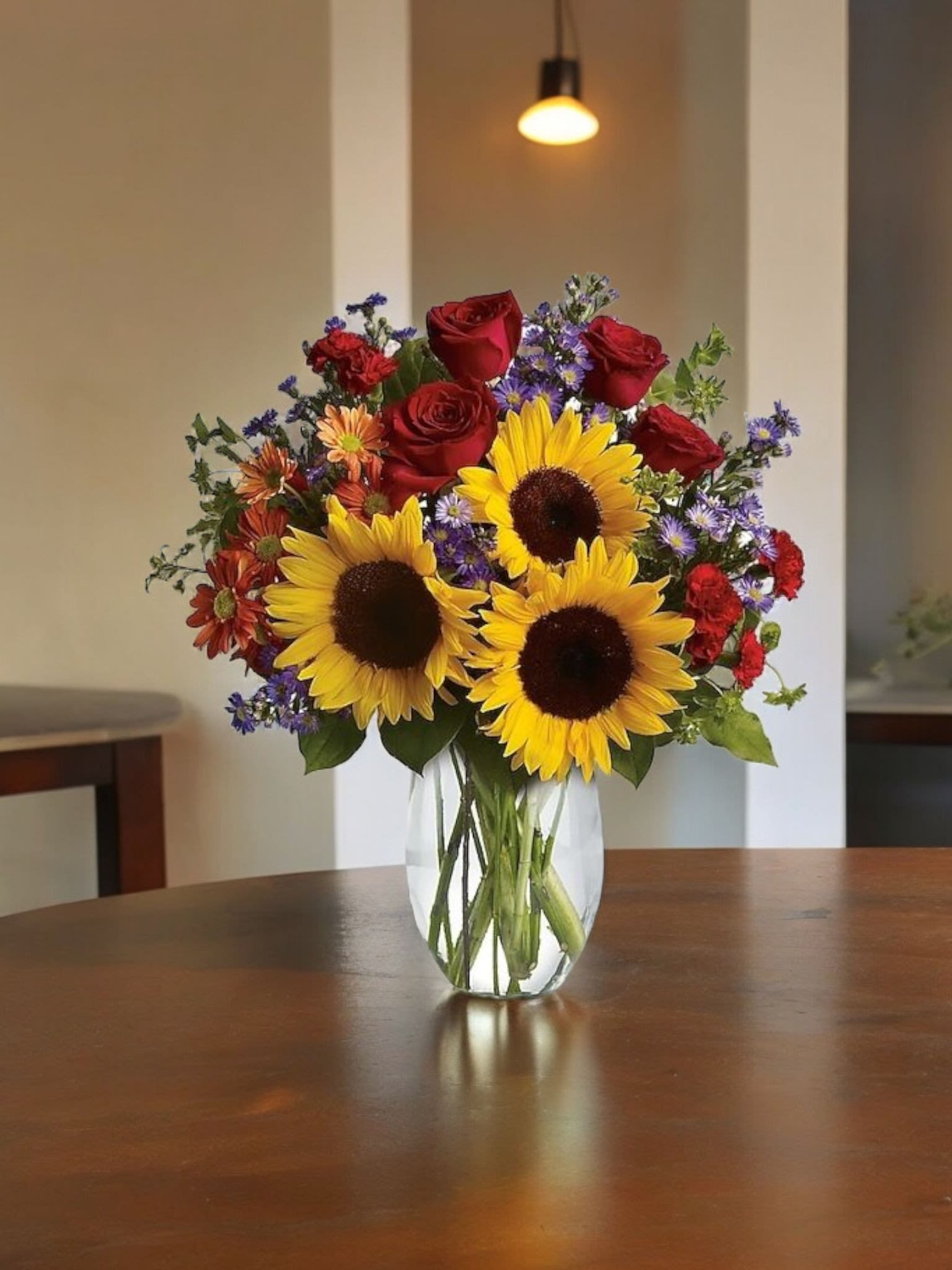 Bouquet of sunflowers, red roses, and purple flowers in a clear glass vase