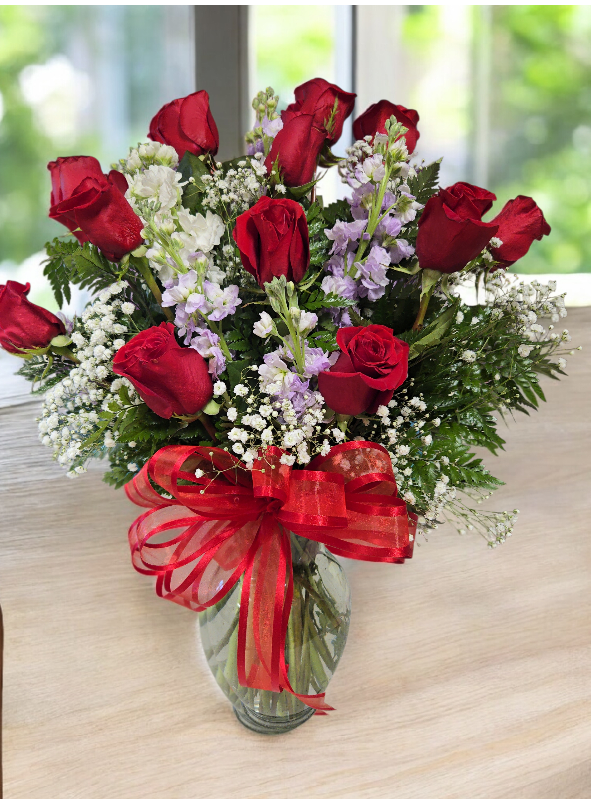  Classic Romance roses -  A timeless bouquet of radiant red roses accented with delicate white baby’s breath, lavender blooms, and lush greenery, presented in a clear glass vase with a bold red ribbon. Perfect for expressing love, admiration, and heartfelt emotions.  
