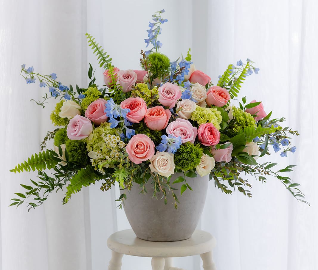 Low gray pot of pink roses, green hydrangeas, and blue delphinium arranged wide on a stool