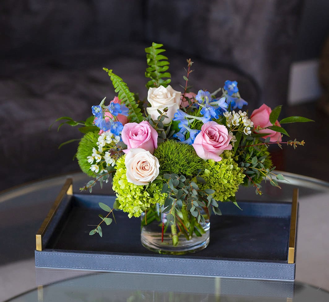 Low arrangement of pink and ivory roses with blue flowers in a clear glass vase