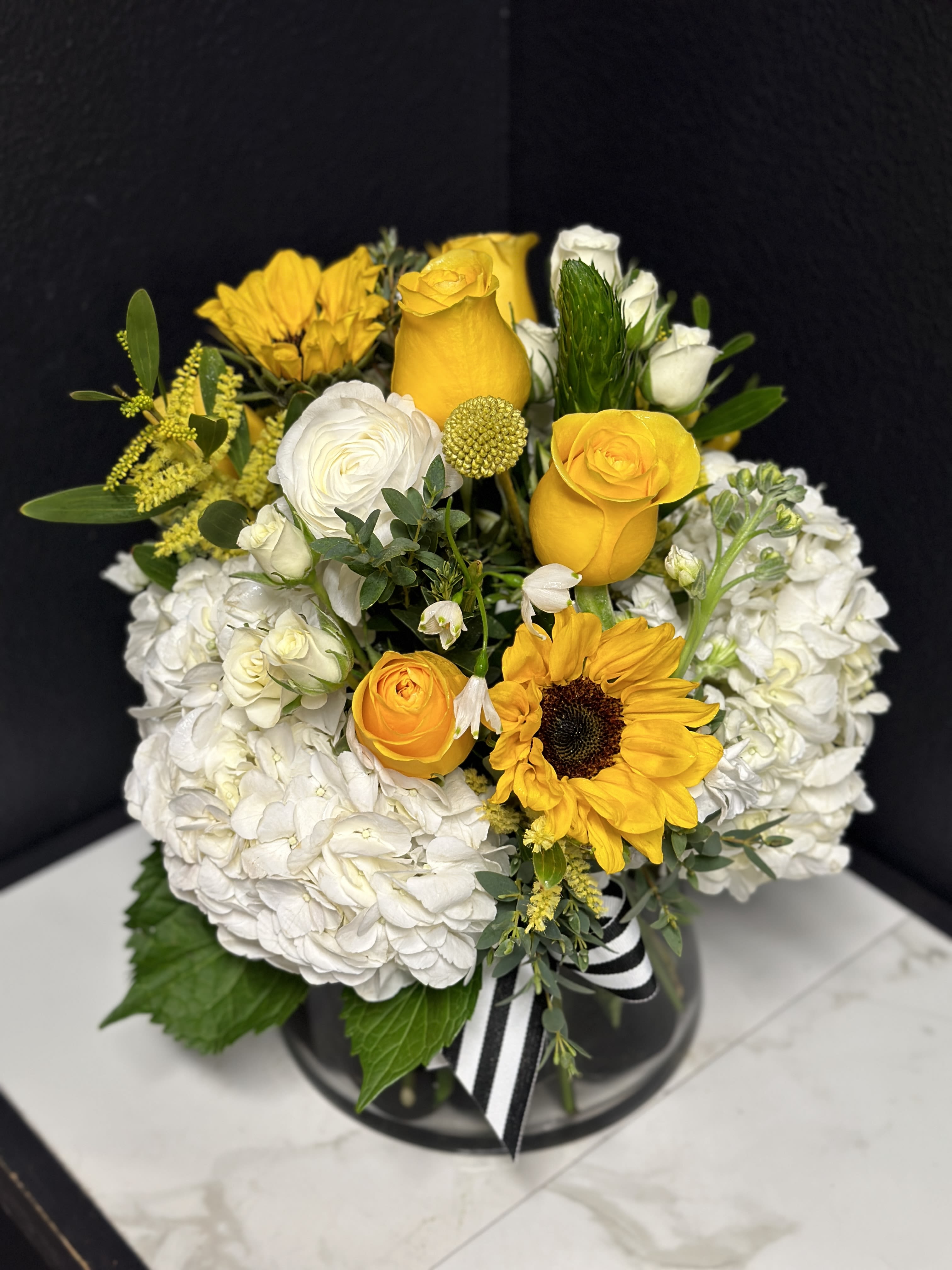 Yellow roses and sunflowers arranged with white hydrangeas in a black vase