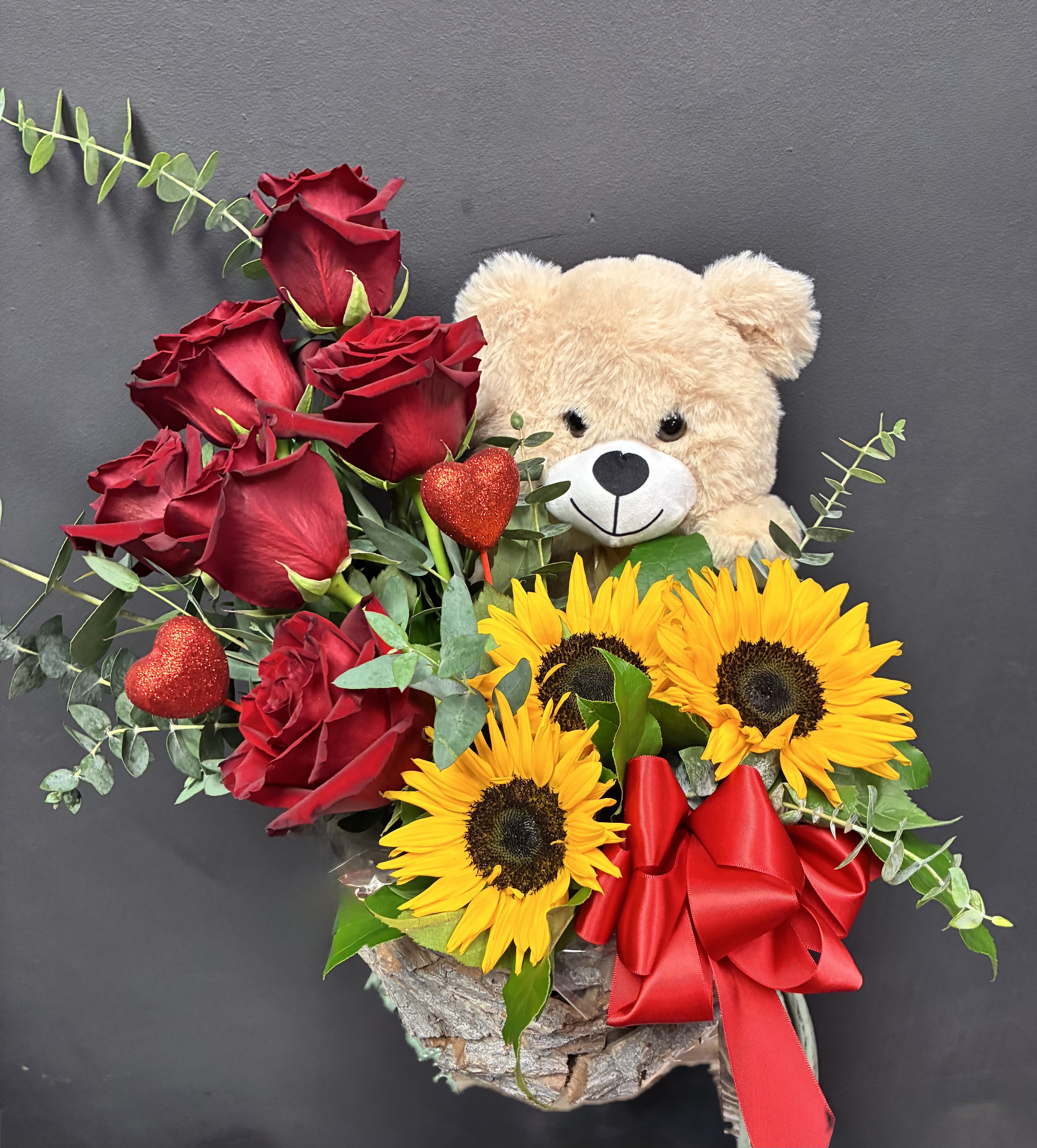 Red roses and sunflowers with a teddy bear and red ribbon in a rustic container