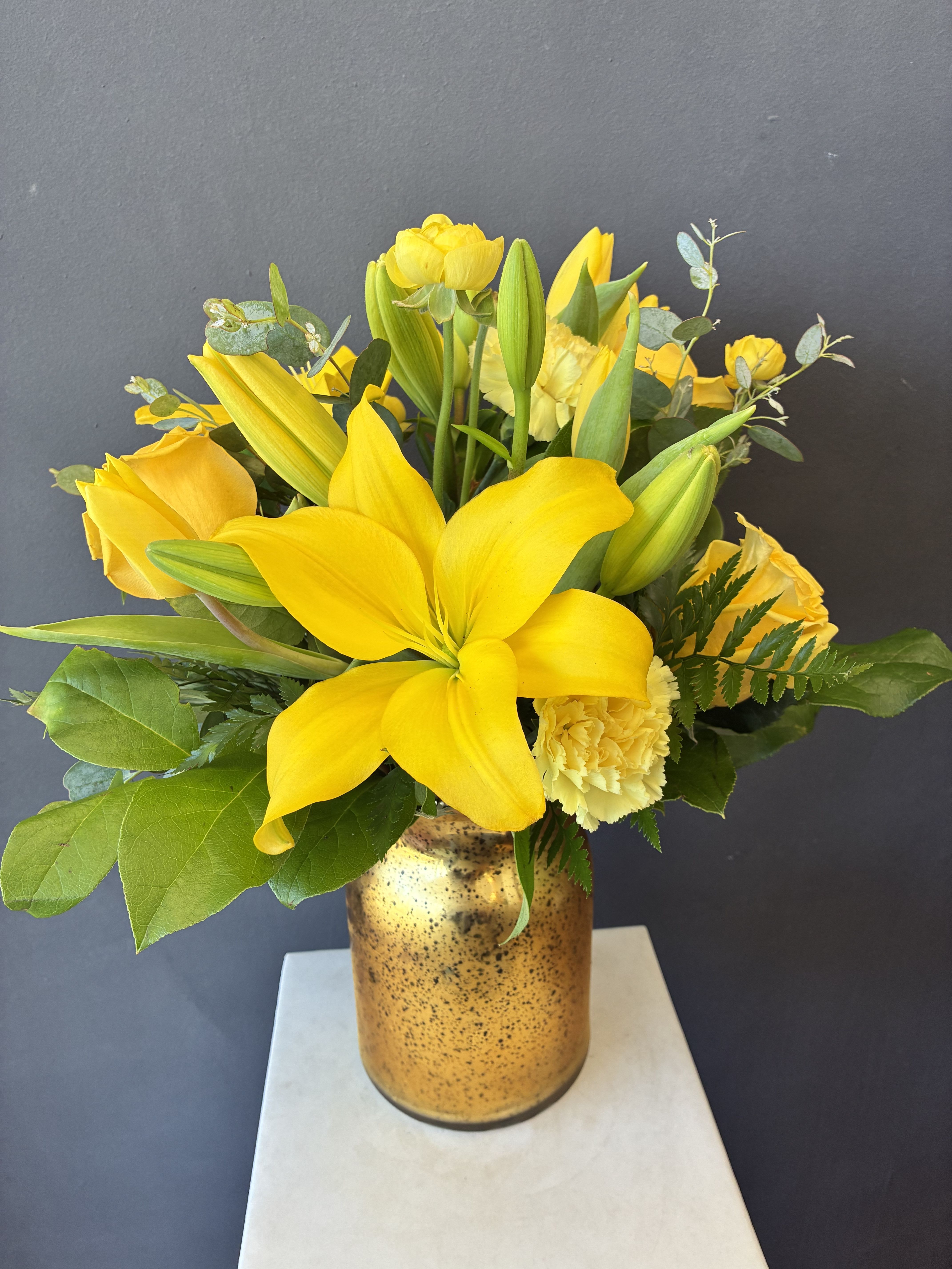 #16 DESIGNERS CHOICE  - Bright yellow Asiatic lilies take center stage in this sunny flower arrangement, a cheerful bouquet perfect for a birthday bouquet or a thoughtful get-well gift that says "cheer up" without words. Framed by yellow tulips and soft yellow carnations, the monochromatic palette reads as joyful and optimistic-ideal for celebrating milestones, offering encouragement, or brightening someone's day.  The stems are gathered in a cylindrical gold-speckled glass vase that adds a warm metallic glow and a modern touch. A mix of open blooms and fresh buds creates movement and a just-picked freshness in a compact, upright shape that fits tabletops and desks. Choose the size that suits your moment and enjoy simple care: fresh water and a cool spot will keep the lilies, tulips, and carnations looking lively for days.