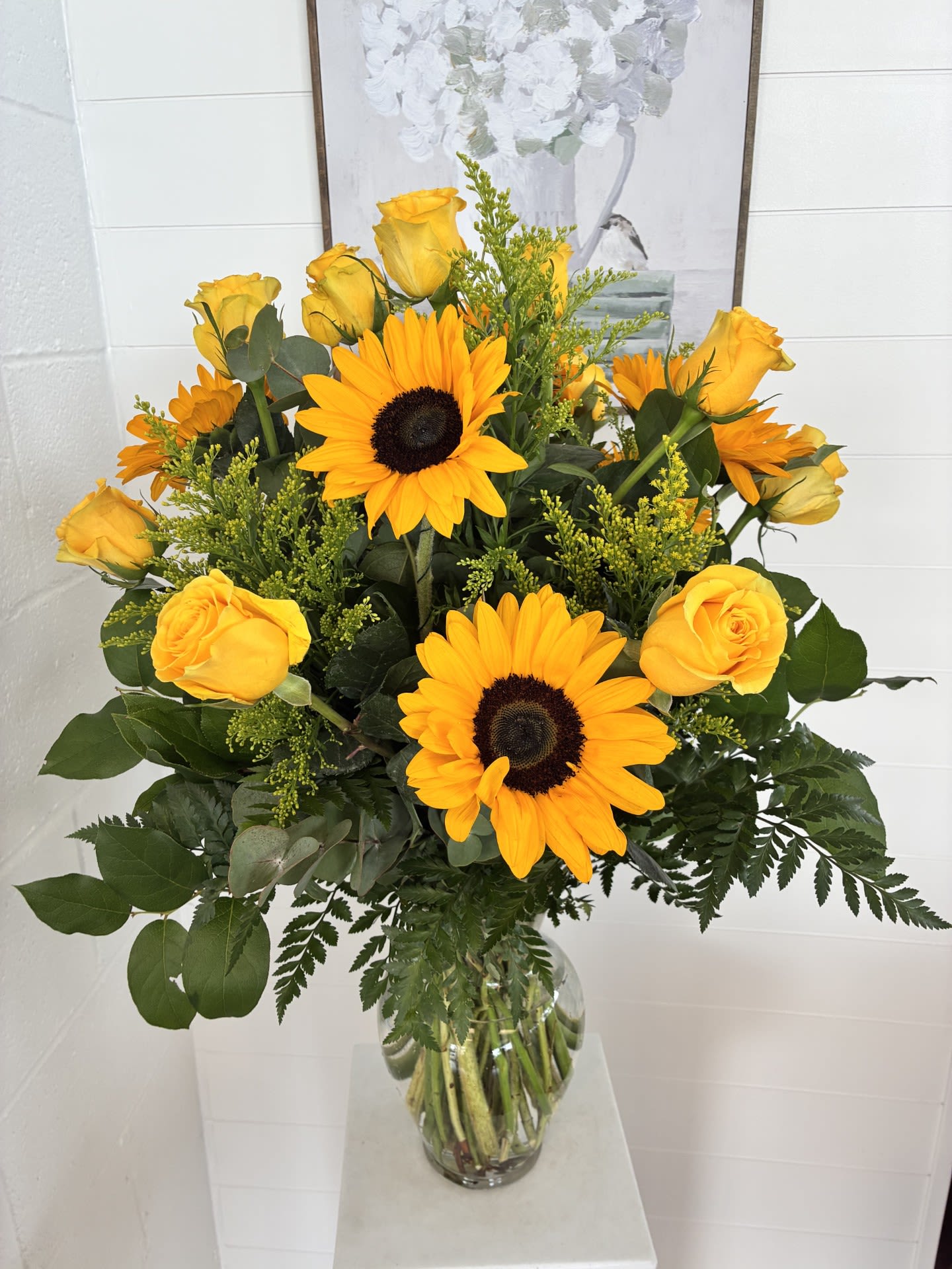 #22 - Bright yellow sunflowers and yellow roses create an immediate burst of sunshine in this flower arrangement-perfect as a birthday bouquet, a thank-you gesture, or a cheerful everyday gift to brighten someone's day. The sunny combination and full, rounded silhouette say "you matter" in a bold, joyful way that will make the recipient feel genuinely seen and uplifted.  The arrangement pairs large sunflowers with warm yellow roses, small yellow filler blooms, and fresh greenery for texture, all gathered in a clear glass vase that showcases the stems and keeps the look clean and modern. Dark sunflower centers add striking contrast to the warm petals, while the compact, dome-like design makes this piece easy to display on a table, desk, or mantel. Sized for easy placement, it arrives ready to enjoy-just add water and let the vibrant yellow and green palette bring a little sunshine into their room.