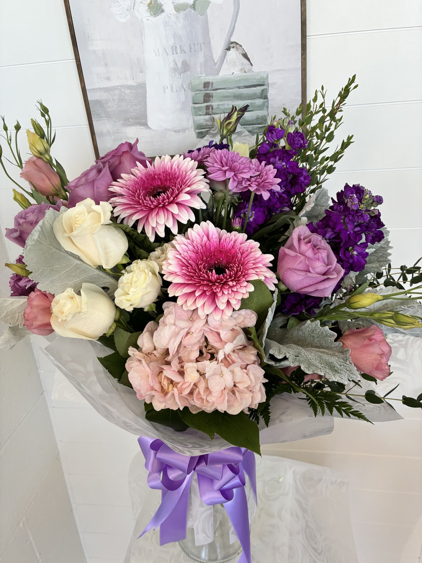 Soft, With Love - Soft, With Love greets her with a lavish garden-fresh embrace-an elegant flower arrangement perfect for a birthday bouquet, anniversary gift, Mother's Day surprise, or any moment you want her to feel cherished. The combination of large pink gerbera daisies and lavender-cream roses makes a romantic first impression, while the pale pink hydrangea and clusters of purple stock add soft volume and depth so the recipient will feel truly seen.  This bouquet mixes roses, gerbera daisies, hydrangeas, lisianthus, and stock in a palette of pink, purple, white, and green for a lush, layered look. Silver-toned foliage and pale lisianthus highlights give subtle contrast; the stems are presented in a clear glass vase with clear plastic wrap and finished with a purple satin ribbon for a polished, gift-ready presentation. The arrangement reads feminine and cheerful without being fussy-textural blooms and soft hues combine for an image-rich, tactile bouquet.  Ideal for the person who deserves warmth and celebration, this arrangement sends a message of affection and appreciation that feels both intimate and special. Dress it up for anniversaries or keep it casual for a "thinking of you" delivery; ordering is straightforward and the vase makes it ready to display on arrival with minimal care-fresh water and a cool spot will help keep the blooms looking their best.