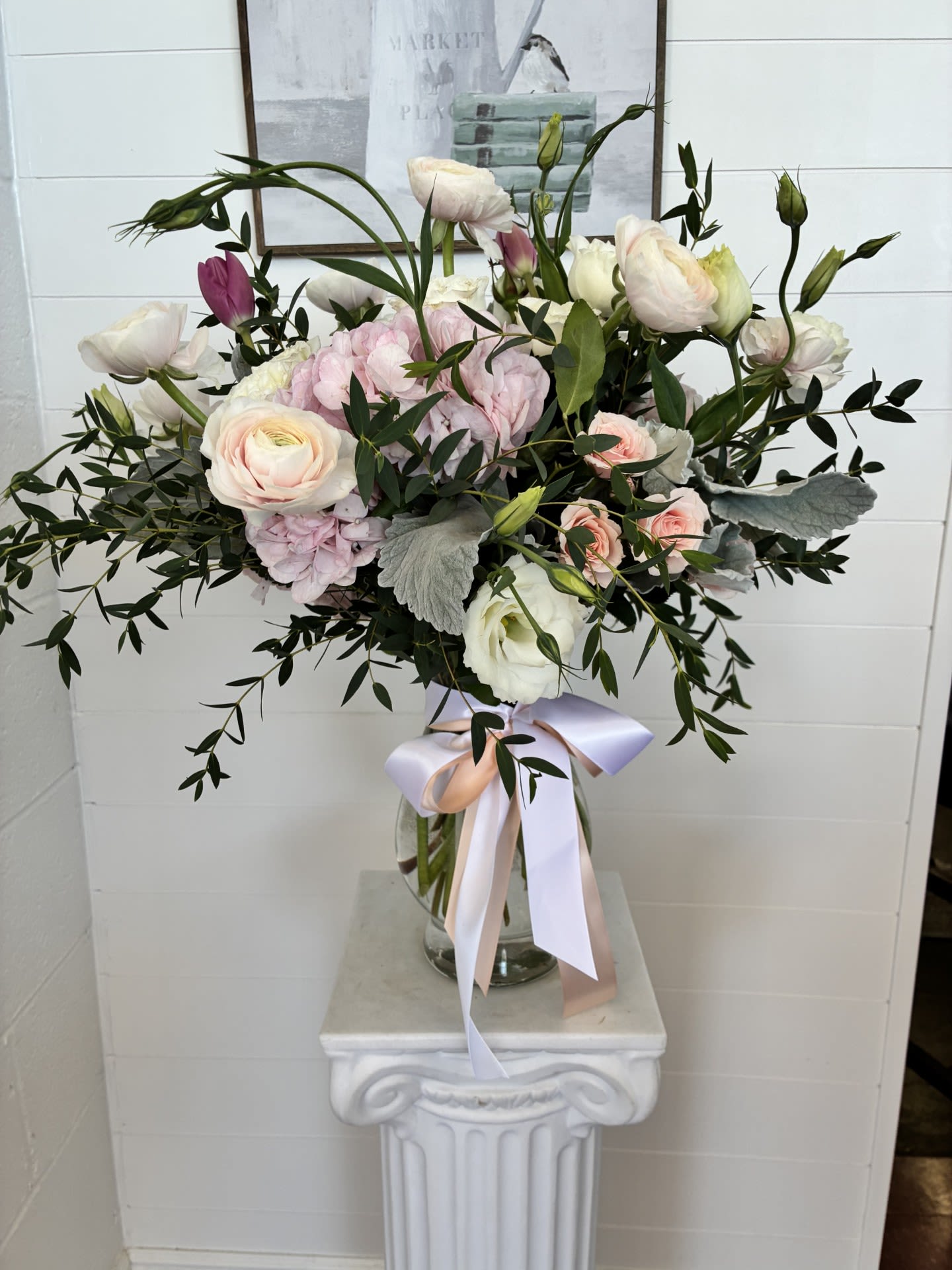 Tender Moments - Pale pink and white blooms mingle with soft mauve accents in this romantic flower arrangement-perfect as a wedding centerpiece, anniversary bouquet, sympathy tribute, or a thoughtful everyday gift. The gentle color story and airy silhouette create a calm, reassuring presence that will make the person you're thinking of feel comforted and truly seen.  Creamy ranunculus-like blooms pair with clustered hydrangea heads, delicate lisianthus, and roses for layered texture and a lush, garden-fresh look. Dark foliage and a few long, arching stems give the arrangement a relaxed, elegant shape. It arrives in a clear glass vase finished with a wide satin ribbon in blush and lavender tones for a refined, hand-tied presentation that reads both polished and personal.  Ideal for someone who appreciates soft, timeless florals, this arrangement suits intimate celebrations, a message of sympathy, or simply brightening a bedside or entry table. Hand-delivered in its vase, it arrives ready to display; refresh water every two days and trim stems as needed to prolong bloom life.