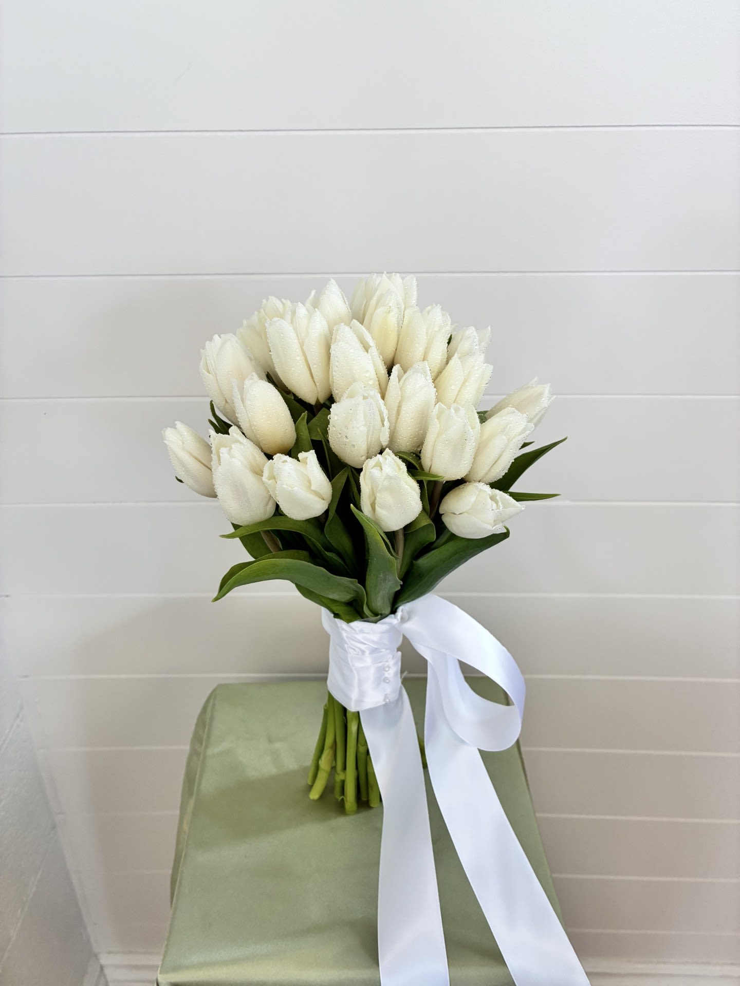 Classic Tulip Wrap - A rounded hand-tied bridal bouquet of white tulips that reads as quietly elegant and fresh - perfect for weddings and formal white-themed celebrations. The Classic Tulip Wrap gives the person you're gifting a clean, timeless look: tightly closed to just-opening tulip blooms create a soft, refined texture that flatters classic bridal styles and minimalist receptions alike.  This wrap features a dense cluster of white tulips nestled against broad green leaves for a crisp two-tone contrast. Stems are bound in white satin ribbon that trails gently for a polished finish and easy carrying; the compact, rounded shape sits comfortably in the hand and photographs beautifully from every angle.  Thoughtful for brides, attendants, or anyone marking a white-themed event, this bouquet sends a message of purity and understated grace - the kind of arrangement that makes someone feel calm, cherished, and perfectly put together on their day. Order with standard sizing suitable for handheld bouquets; stems are ribbon-wrapped for neat presentation and simple care (keep stems in water until ready to use).