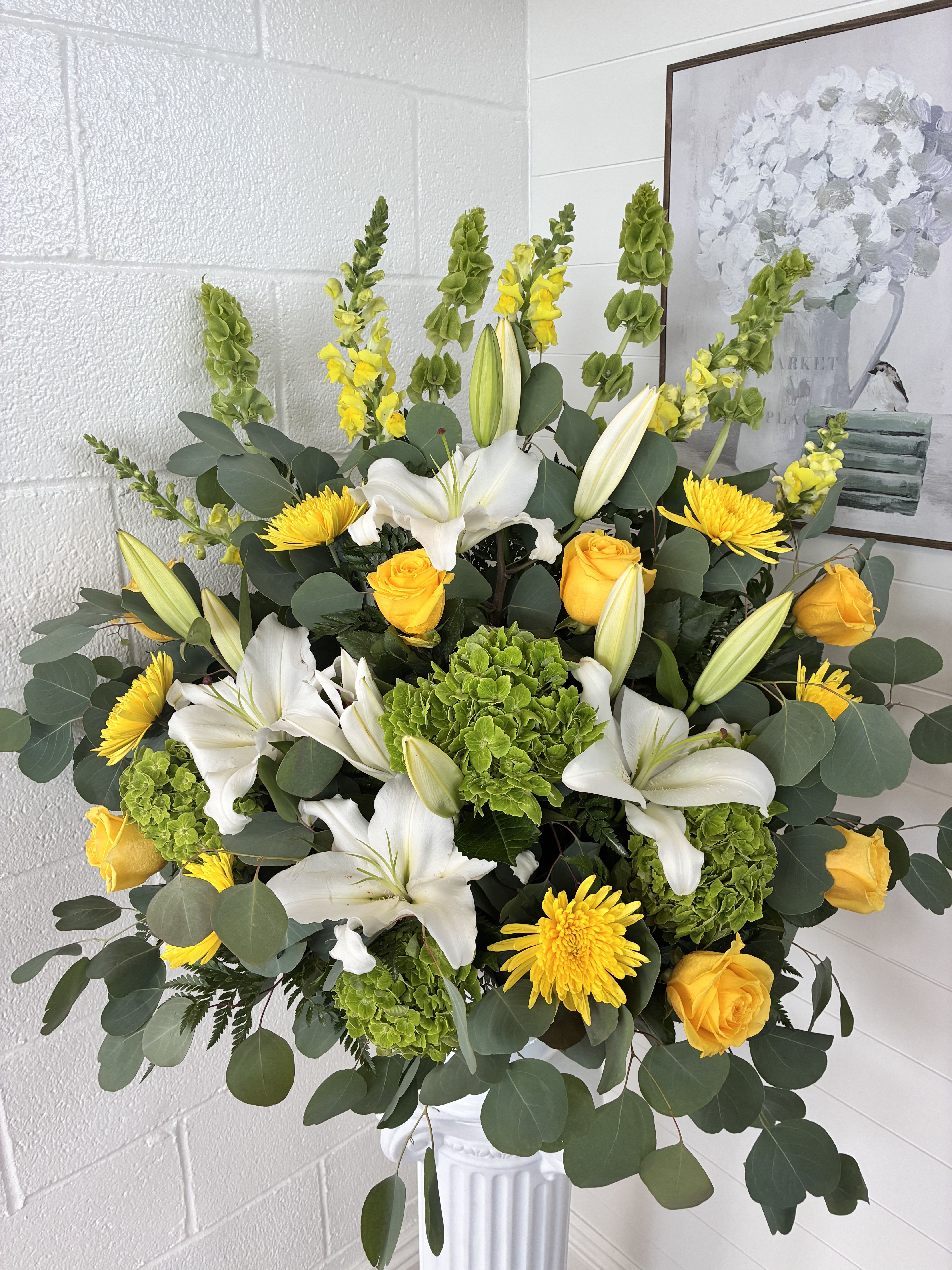 Golden  - Sunlit white lilies and sunny yellow roses create a joyful, uplifting presence in this large upright flower arrangement-perfect as a birthday bouquet, cheerful thank-you gift, or bright centerpiece for spring celebrations. The tall, garden-style silhouette and layered height make it the kind of arrangement that greets the recipient with warmth and effortless elegance, so the person you're thinking of will feel genuinely celebrated.  This design pairs fragrant white lilies with yellow roses, yellow chrysanthemum blooms, and rounded green hydrangea-like clusters set against dense eucalyptus and vertical green spikes for added height. Arranged in a classic white pedestal vase, the palette of white, yellow, and green reads fresh and polished while the varied textures-from the soft lily petals to the rounded green clusters-keep the composition lively and refined.  Ideal for sending to mom, a friend, or a grateful colleague, this arrangement arrives ready to display and makes an immediate, sunny impression on any table or entryway. Ordering is simple-choose delivery to brighten someone's day or pick up for an easy, last-minute gift that looks thoughtfully arranged and professionally finished.