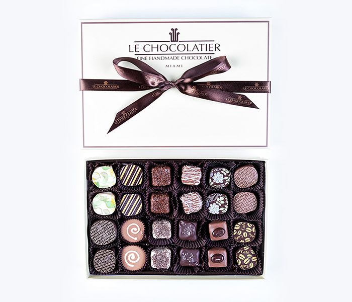Rectangular box of assorted gourmet chocolates with lid and brown ribbon beside open tray