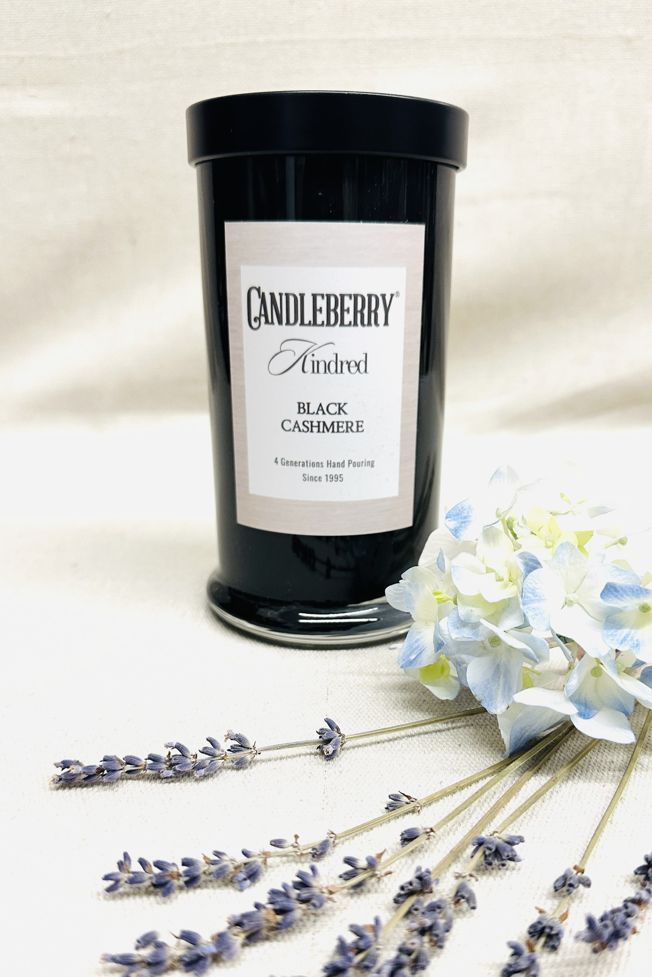 Black candle jar with a label beside pale blue and white flowers