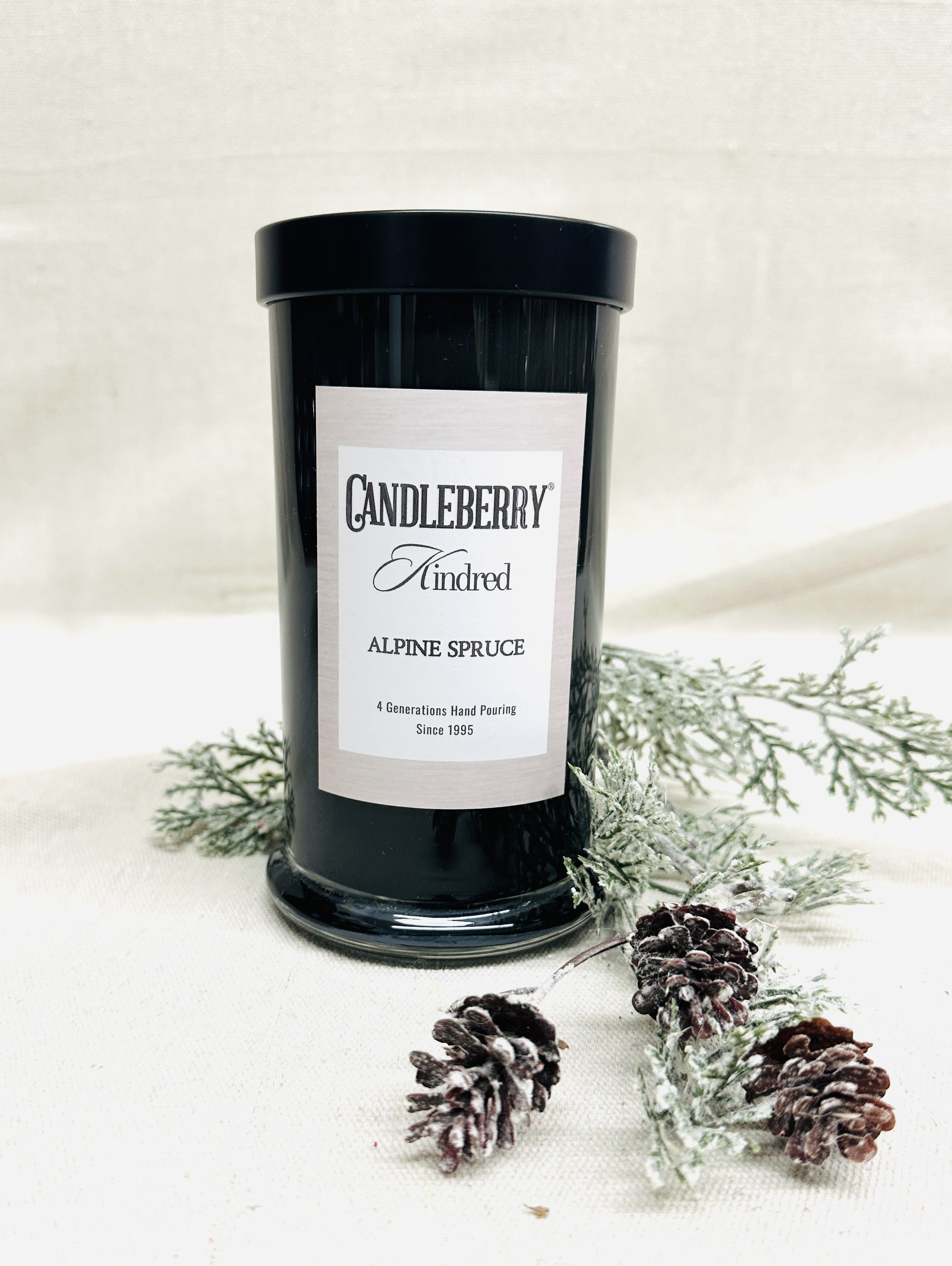 Black candle jar labeled Alpine Spruce with frosted pine sprigs and pinecones