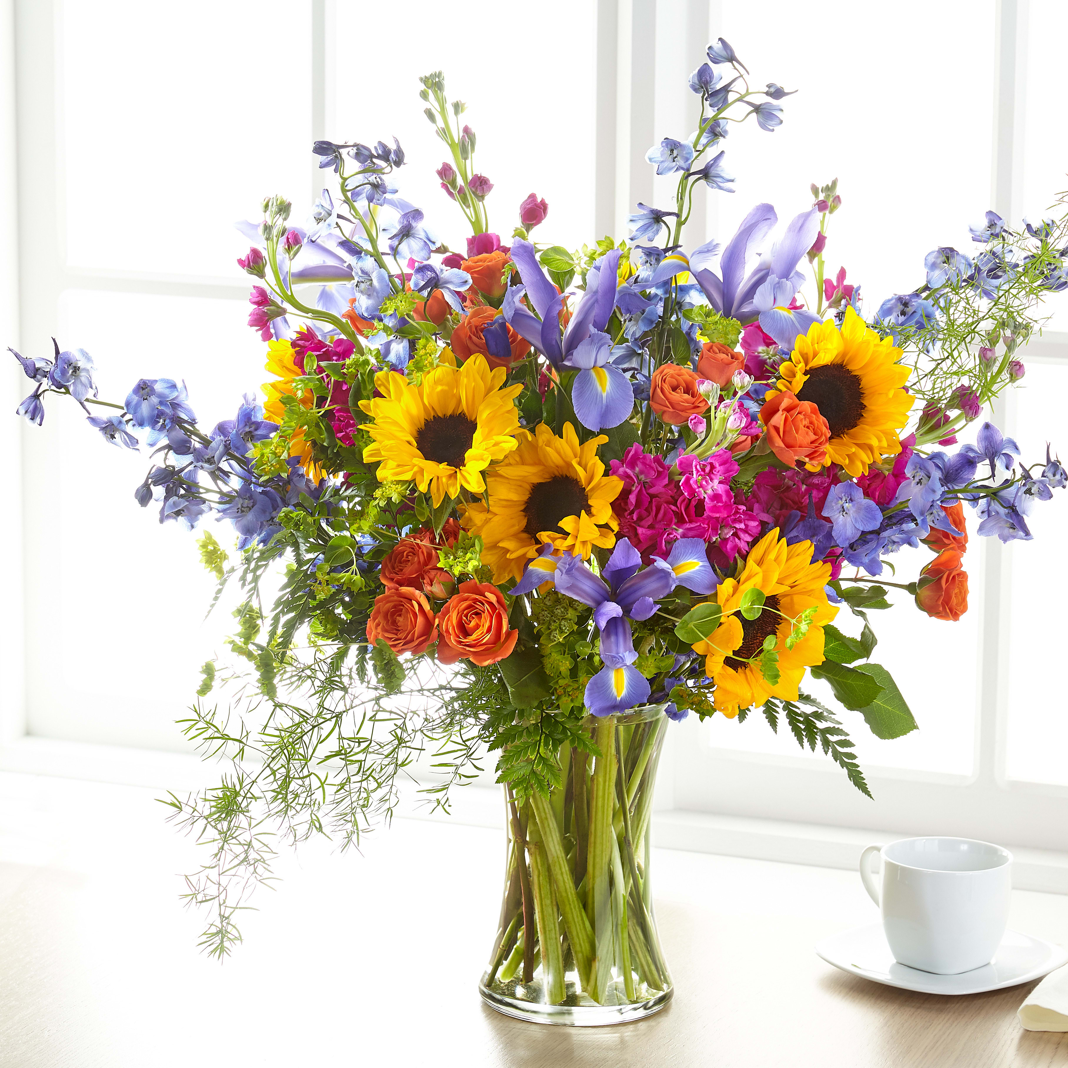 Tall mixed bouquet with sunflowers, blue irises, delphinium and orange roses in a clear glass vase.