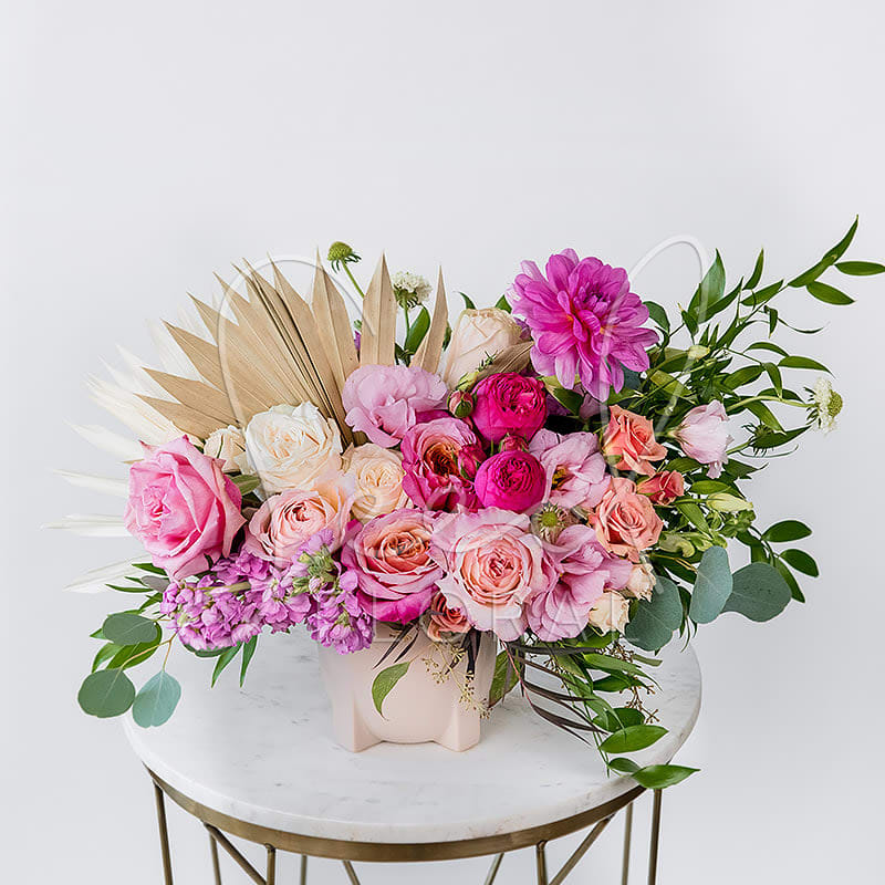The Boho Babe - Free-spirited and full of charm, Boho Babe is a modern floral arrangement with a playful twist. Featuring a stunning mix of garden roses, ranunculus, lisianthus, and stock in romantic shades of pink, peach, cream, and bold pops of magenta, it’s artfully accented with dried palm leaves and lush greenery for that effortlessly chic, boho-luxe vibe. Presented in our signature Modern Smooth Taupe Vase, this arrangement is perfect for making a statement — whether you're celebrating love, saying thank you, or simply treating someone to something unforgettable.