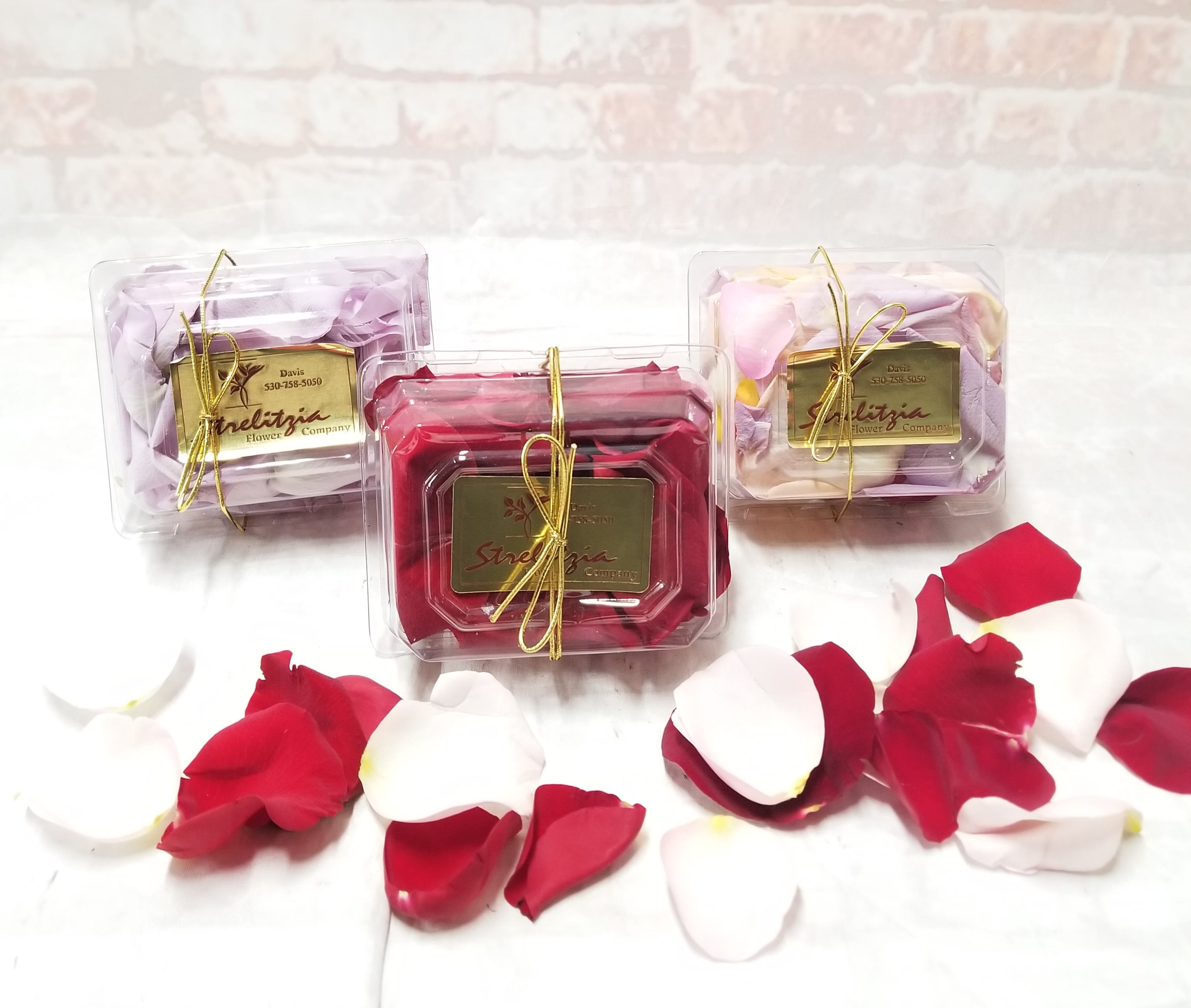 Three clear gift boxes of rose petals tied with gold ribbon.