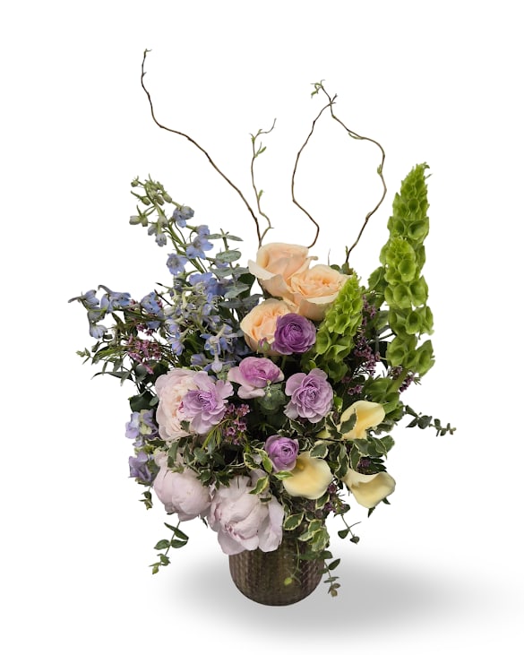 Spring Gem - Soft lavender delphinium and pale yellow calla lilies lift a cascade of mixed-color roses, pink ranunculus, and airy blue Bella Donna into a sculptural, springlike display-perfect as a Mother's Day gift, birthday bouquet, or thoughtful "get well" surprise. The tall curly willow branches give the arrangement dramatic vertical presence while keeping the feel open and romantic, so the person you're thinking of will feel genuinely celebrated and seen.  This floral arrangement includes roses, ranunculus, peony, hydrangea, stock, bells of Ireland, wax flower and calla lilies arranged in a dark, textured keepsake vase with curly branches for height. Dimensions are approximately 26" high by 17" wide; arrange in a stable spot and refresh water every other day, recutting stems if needed to extend vase life.