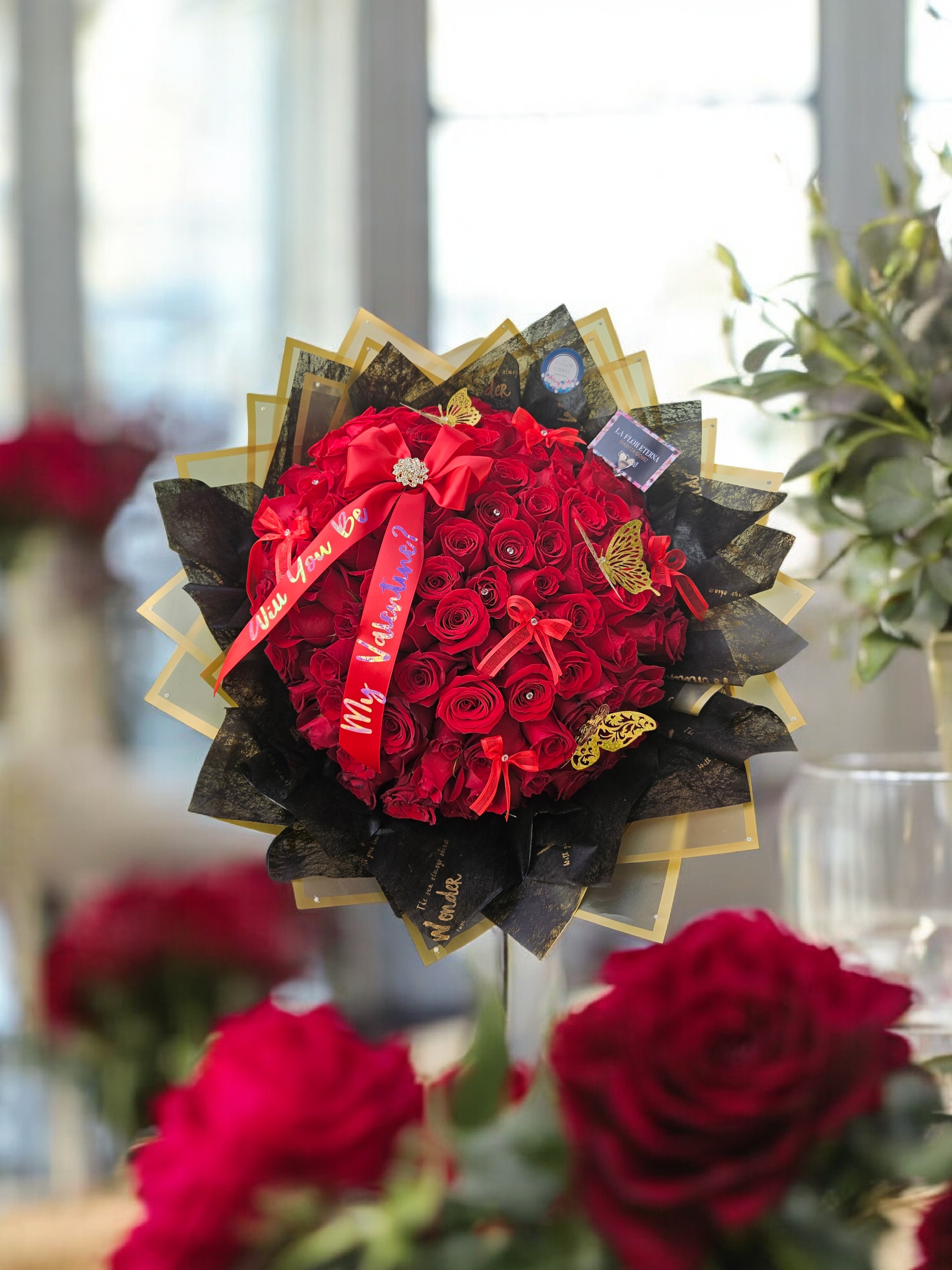 Round bouquet of tightly packed red roses wrapped in black and gold paper with red ribbons.