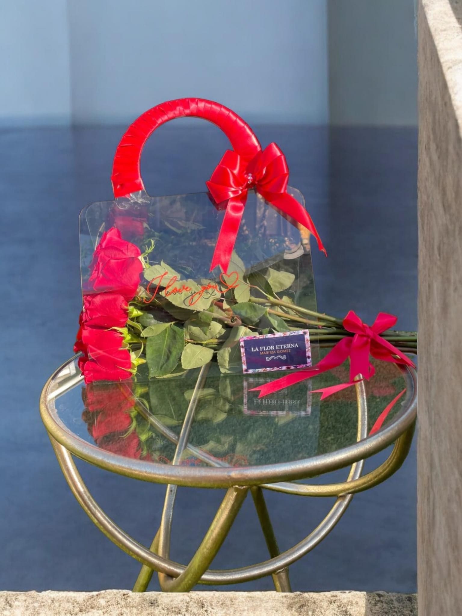 Love Purse - A small bundle of long-stem red roses arranged for a modern romantic moment - perfect as a birthday bouquet, anniversary roses, or a simple "thinking of you" Valentine's gift. The clear plastic handbag-style gift bag with its bright red wrapped handle and satin bow gives the presentation a fresh, contemporary feel so the person you're celebrating arrives to an immediate, confident impression of love and attention.  The roses are tied with a matching red satin ribbon and come with a printed gift card bearing the florist's branding, displayed here on a round glass-top metal table to show scale and styling. The compact, handheld size makes this an intimate, easy-to-carry gift that reads as thoughtful and striking; choose it when you want her to feel noticed and cherished without fuss.