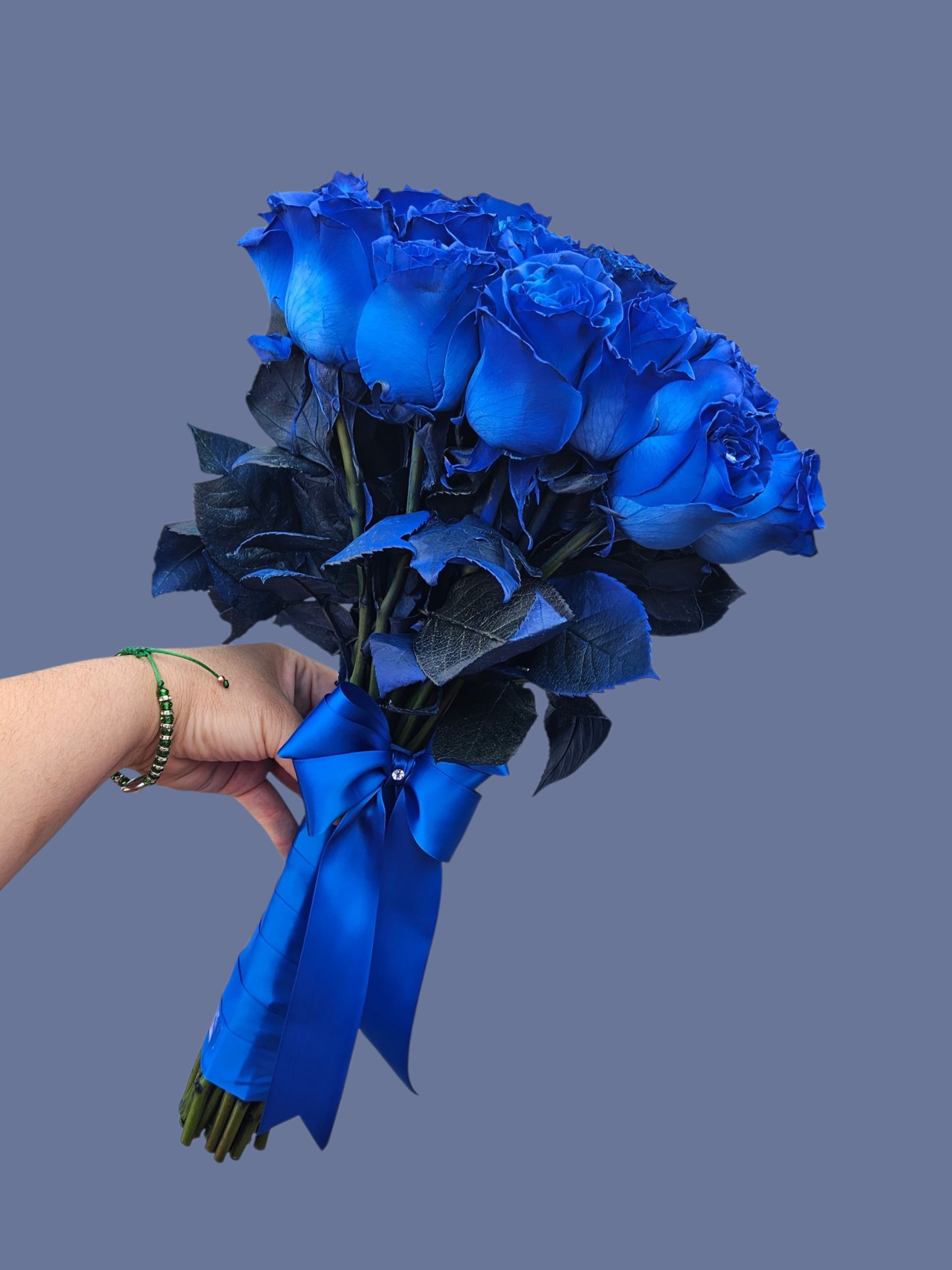 Nake blue  - A dense cluster of vivid blue roses creates an immediate, dramatic impression-this handheld flower arrangement is built for romance and bold celebrations. As a blue rose bouquet for anniversaries, birthdays, or a congratulatory gift, it reads as both intimate and formal: the saturated petals draw the eye while the dark foliage and stems sharpen the contrast so the blooms feel jewel-like.  Tightly hand-tied in a classic compact shape, the bouquet is finished with a wide matching blue satin ribbon and long tails for a polished presentation. The deep blue and black-toned foliage gives the arrangement a slightly edgy, sophisticated mood that will make the person you're thinking of feel truly seen and celebrated.