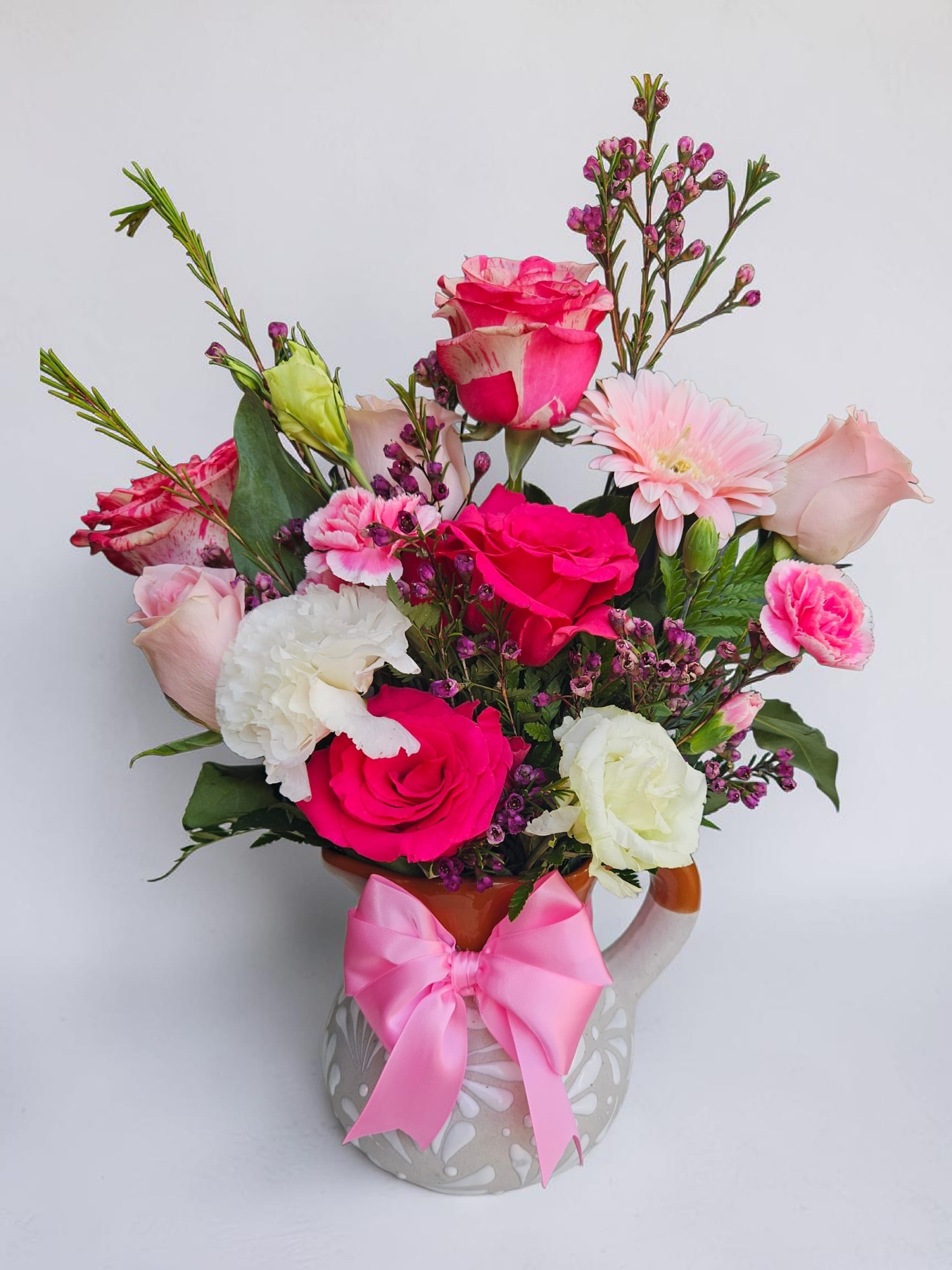 Mexican Jara  - Bright, playful color takes center stage in this mixed flower arrangement-perfect as a birthday bouquet, Mother's Day gift, or an everyday just-because surprise. Hot pink and blush roses pair with white and pale-pink carnations and lisianthus-style daisy blooms to create a romantic yet energetic look, while pops of purple filler and sunny yellow sunflowers or gerbera accents lift the palette into cheerful territory. The overall effect reads feminine and joyful, the kind of bouquet that makes the person you're thinking of feel noticed and celebrated.  Arranged compactly in a decorative ceramic mug or rounded ceramic vase and finished with a large pink satin ribbon bow, this design is ready to give-no extra wrapping needed. Expect a full, rounded composition of roses, carnations, gerbera-like daisies, small purple filler blooms, and occasional sunflowers for bright contrast; colors include hot pink, blush, white, purple and yellow against fresh green foliage. Sized for countertop display, it's an easy-care, giftable arrangement that brightens a kitchen table, office desk, or bedside table-ideal for birthdays, thank-you gestures, anniversaries, or springtime celebrations.