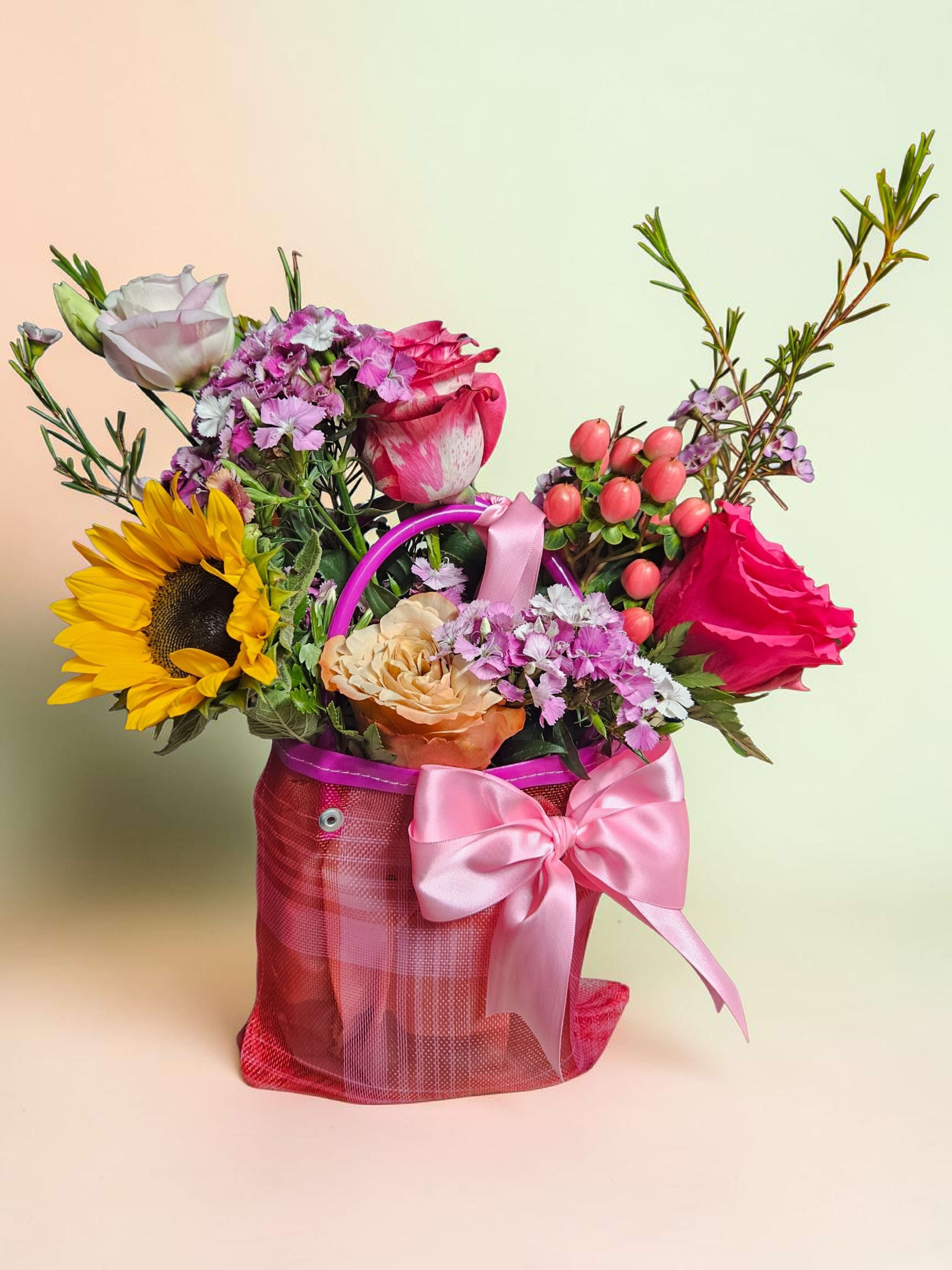 Mini Mexican flower bag  - A bright sunflower crowns this Mini Mexican flower bag, making it a joyful everyday flower arrangement perfect for birthdays, thank-you gifts, or a cheerful "just because" surprise. The mix of pink, peach, white and purple blooms reads both lively and soft-exactly the kind of gift that makes the person you're thinking of feel noticed and celebrated.  Inside the pink gift bag you'll find a tight gathering of sunflowers, roses in pink, white and peach tones, carnations in pink and purple, and clusters of small filler blooms for texture and movement. The presentation includes a pink satin ribbon bow, a purple handle, and a glossy pink mesh wrap that keeps the bouquet neat and gift-ready-small enough to tuck into a desk or bedside table yet visually full and abundant.  This compact arrangement suits many recipients: a friend, coworker, or loved one who appreciates bright color with a romantic, playful touch. Order with confidence for same-week giving; the satin-wrapped bag and secure handle make it easy to deliver this little celebration by hand.