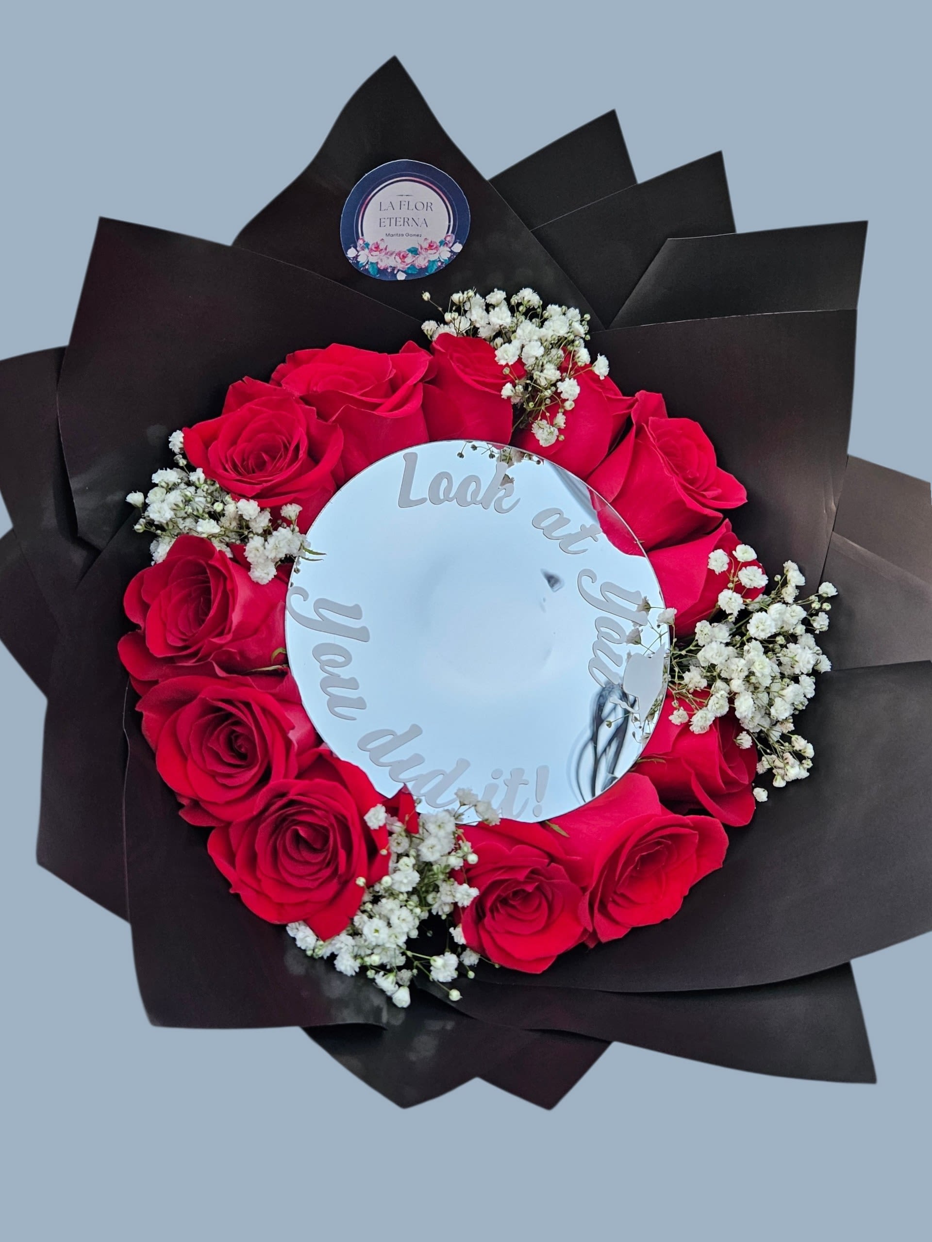 Look at you graduation bouquet  - A bold ring of vivid red roses framed by delicate white baby's breath-this graduation bouquet makes a confident, celebratory statement the moment they walk in. Designed as a circular arrangement with a mirrored center that reads "Look at you, you did it!", it's ideal as a graduation bouquet or a congratulatory gift that tells the recipient they're seen and celebrated.  The bouquet features tightly clustered red roses set around a round mirror disc, with small white filler blooms tucked between petals for contrast and texture. Layers of black wrapping paper and a black floral wrap create dramatic negative space, while a red ribbon and clear decorative pick add polished finishing touches. A printed sticker label and subtle decorative lettering on the mirror give it a personalized, festive feel.  Perfect for the new grad, recent achievement, or milestone celebration, this arrangement feels both romantic and triumphantly modern-the kind of gift that makes someone feel proud and noticed. Sizes and specific stem counts vary by selection at checkout; to keep the mirror bright, avoid prolonged direct sunlight and refresh the water if stems are provided with a vase option.