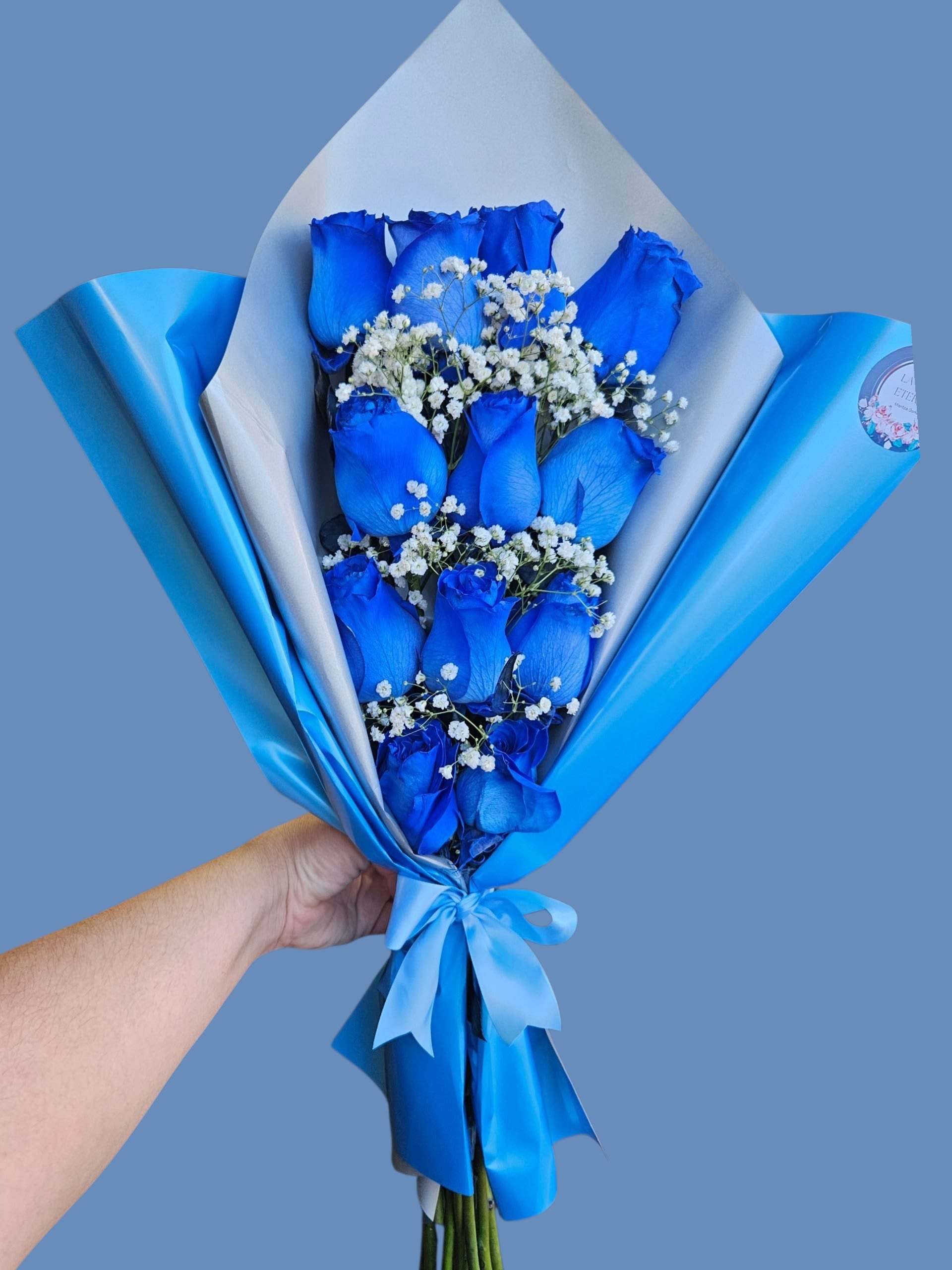The Azure Elegance - Vivid blue roses take center stage in this blue-and-white hand-tied bouquet, a modern romantic choice for an anniversary, birthday, or simply to make someone feel unforgettable. The striking monochrome palette is softened by sprays of white baby's breath, so the person you're gifting will notice both the bold color and the delicate, airy contrast.  Arranged in a compact cluster and wrapped in glossy blue paper with a crisp white inner layer, the bouquet is finished with a matching blue satin ribbon for a polished presentation. Compact and easy to hold, it reads as both celebratory and distinctive-perfect for someone who loves contemporary style with a romantic twist.  Choose this arrangement when you want the recipient to feel noticed and celebrated; it's the kind of bouquet that makes a statement without shouting. Intended as a ready-to-give hand-tied bouquet, no special care is needed beyond fresh water once placed in a vase.