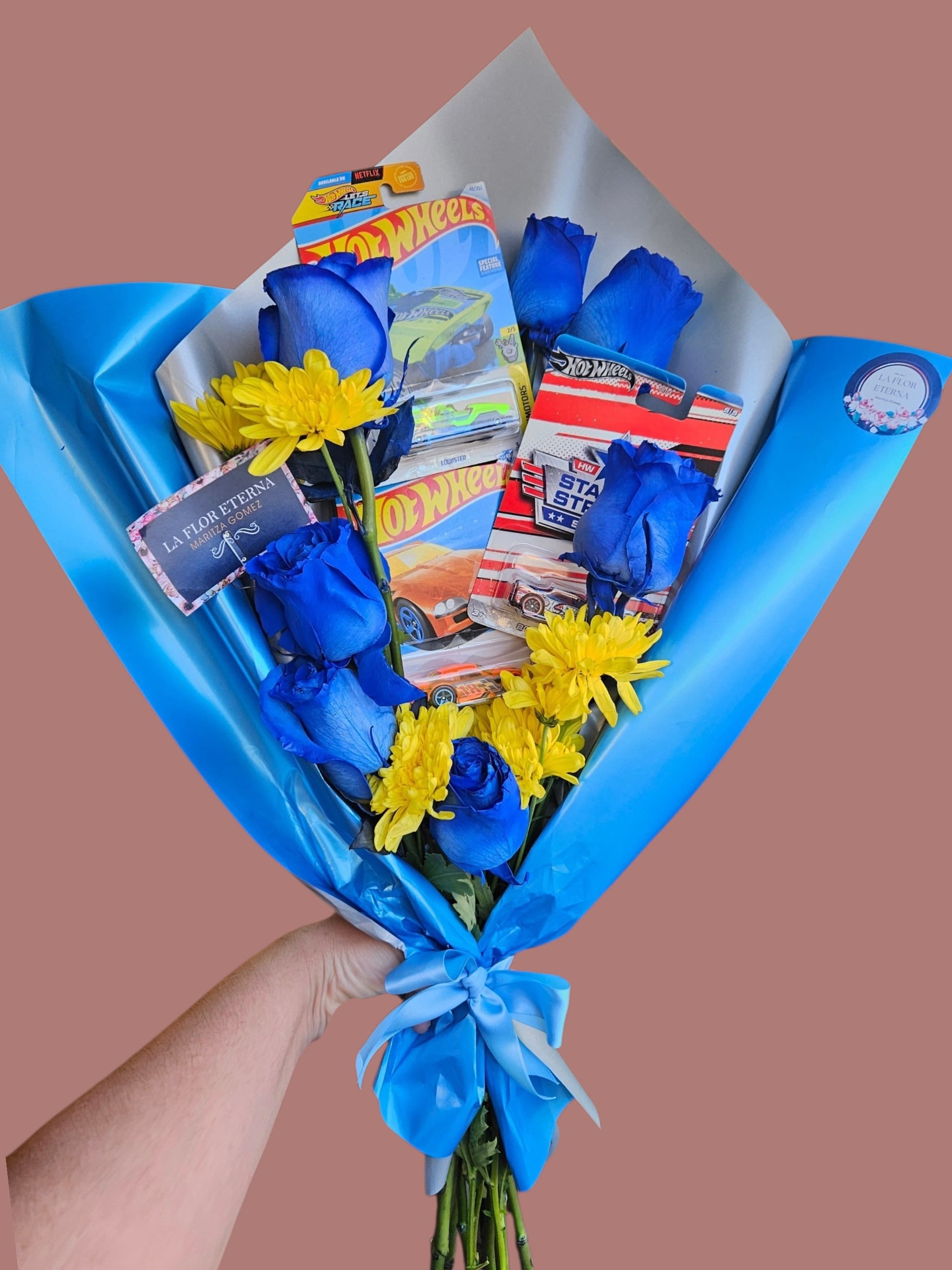 Race Blue Bouquet  - Vivid blue roses and sunny yellow daisies come together in a playful birthday bouquet that feels like a celebration the moment it's handed over. This flower arrangement for birthdays and kid-friendly celebrations pairs striking blue-and-yellow contrast with a whimsical touch-several small toy cars in blister packs nestle among the blooms, so the recipient will feel delighted and seen, whether they're turning five or fifty.  The mix includes richly colored blue roses and bright daisy-like yellow blooms arranged tightly and wrapped in glossy blue decorative paper with a pale inner layer. A satin ribbon finishes the presentation, keeping the look polished and gift-ready. The bouquet's compact, handheld size and the secure wrapping make it easy to carry straight to the party.  Ideal for a child's birthday, a young-at-heart friend, or anyone who loves cars and bold color, this arrangement sends an unmistakable message of fun and celebration. Order with standard handling; keep the flowers in fresh water once unwrapped and place away from direct heat to preserve color and freshness.